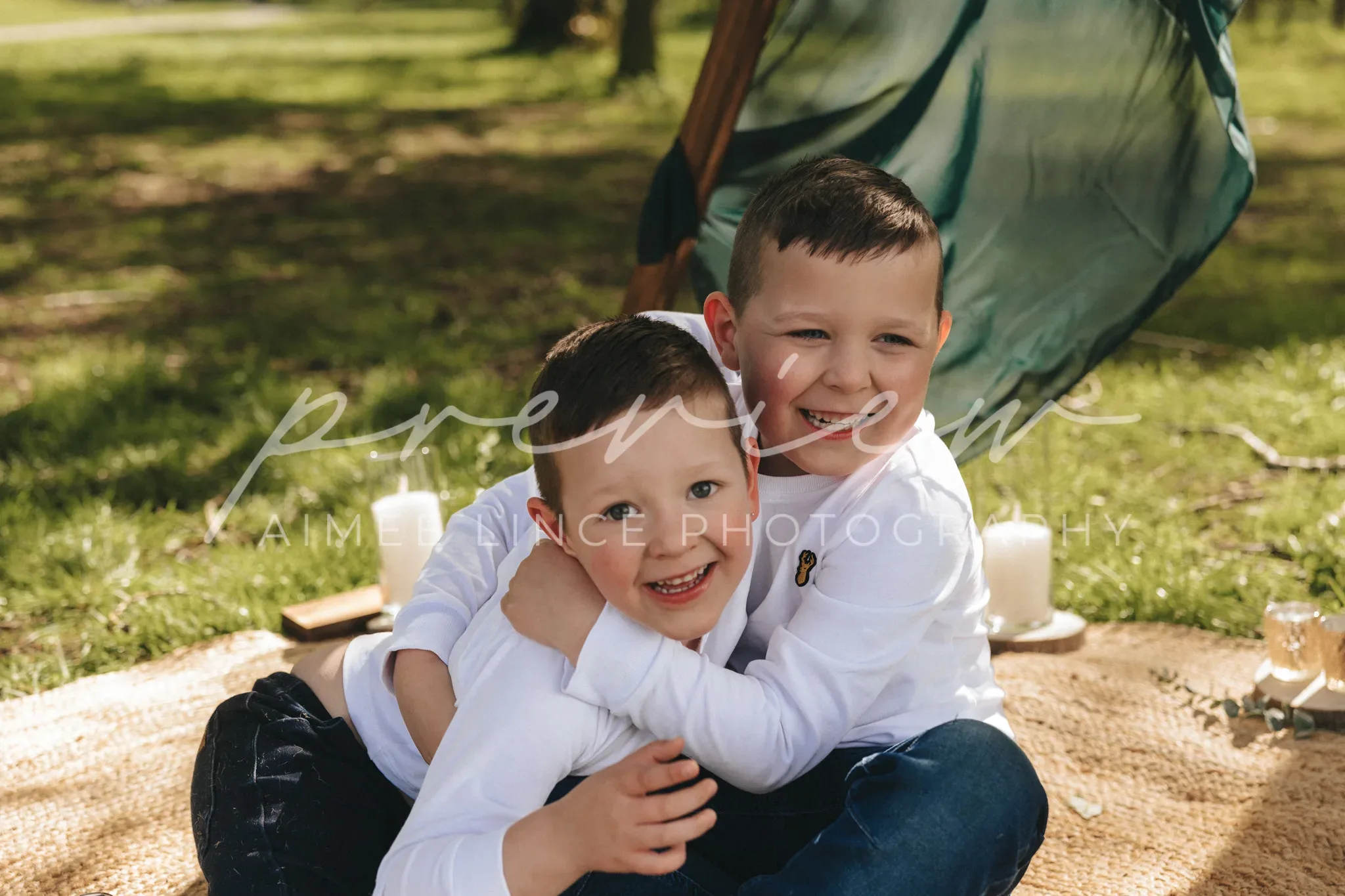 Two young boys smile joyfully as they embrace each other sitting on a blanket in a sunlit park, with a tent and trees in the background. the photo has a watermark labeled "preview, aimée lence photography".