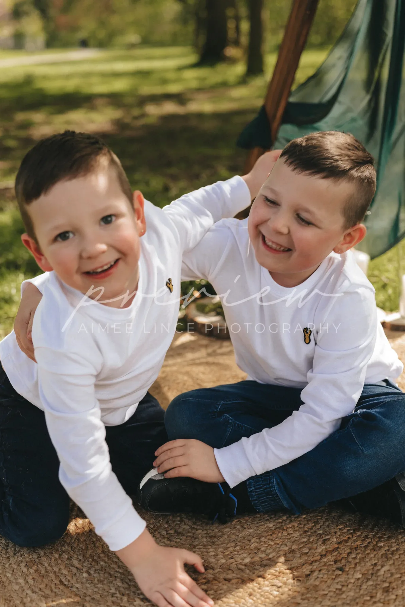 Two young boys, smiling and sitting on a burlap blanket in a sunny park, playfully interact as one reaches out to gently touch the other's ear. both are dressed in white shirts with a small bee emblem.