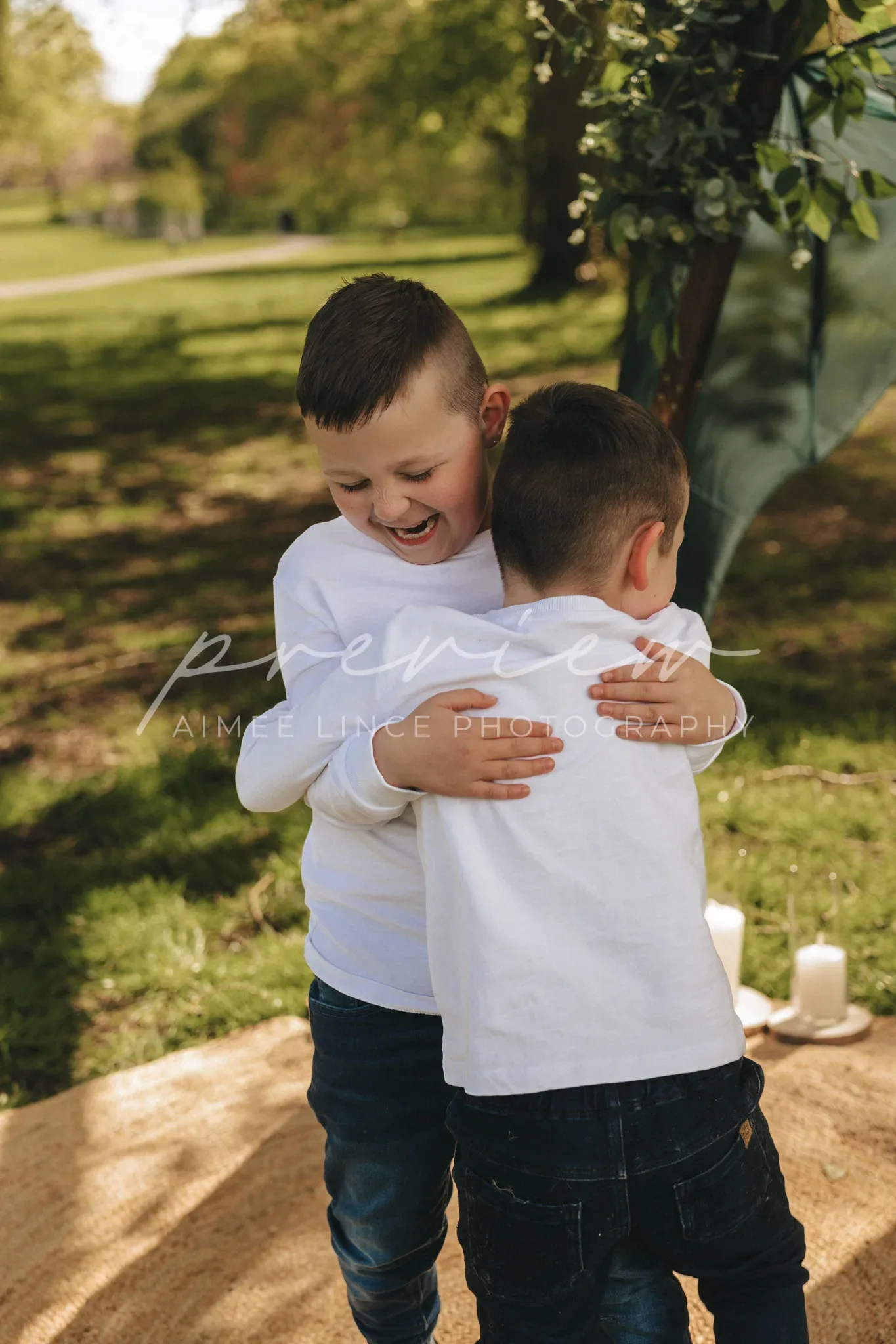 Two young boys hug joyfully in a park. the older boy, wearing a white shirt, smiles broadly as he embraces the younger boy in a blue shirt, whose back is to the camera. sunlight filters through trees in the background, enhancing a warm, cheerful atmosphere.