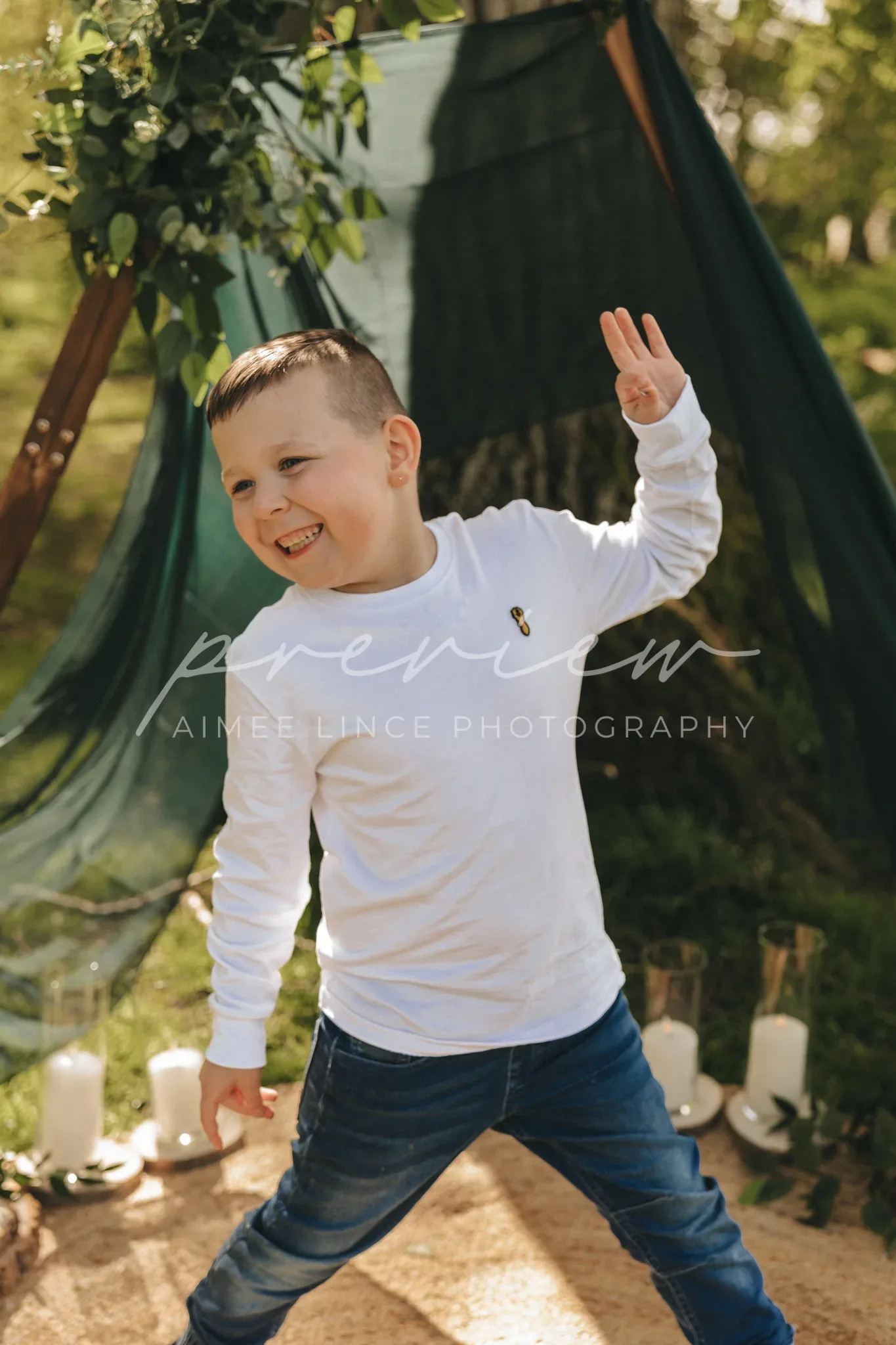 A smiling young boy with short hair throws a peace sign, standing in a garden. he wears a white sweater and blue jeans. behind him, a green fabric teepee adorned with vines adds whimsy to the sunny backdrop.