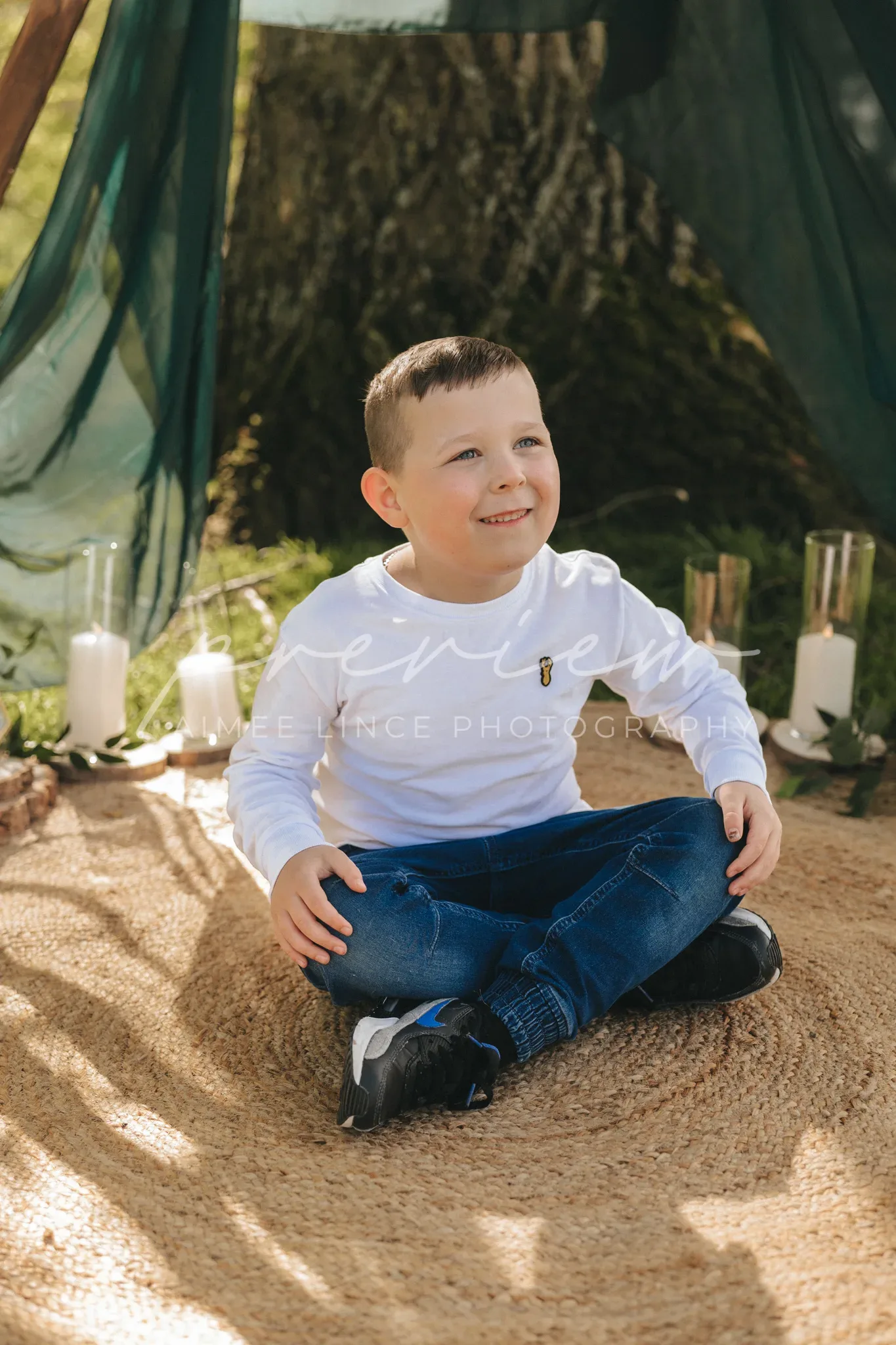 A young boy smiles while sitting cross-legged on a beige rug outdoors, surrounded by green drapes and white candles. he wears a white shirt, blue jeans, and black shoes. the sunlight filters through trees, creating a warm ambiance.