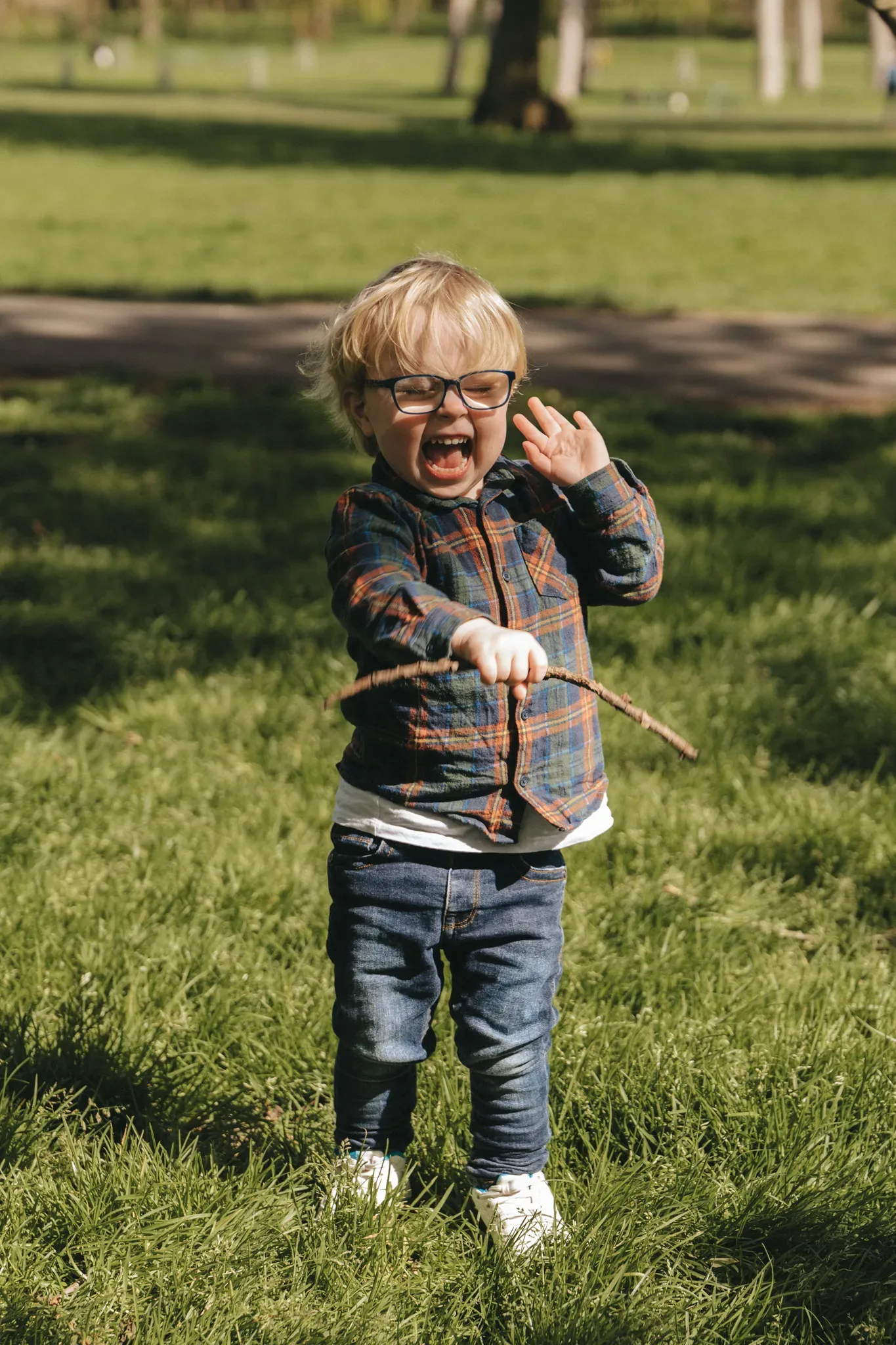 A joyful young boy with blond hair and glasses laughs while holding a stick in a park. he wears a plaid shirt, jeans, and sneakers. the background shows lush green grass and scattered trees under a clear sky.