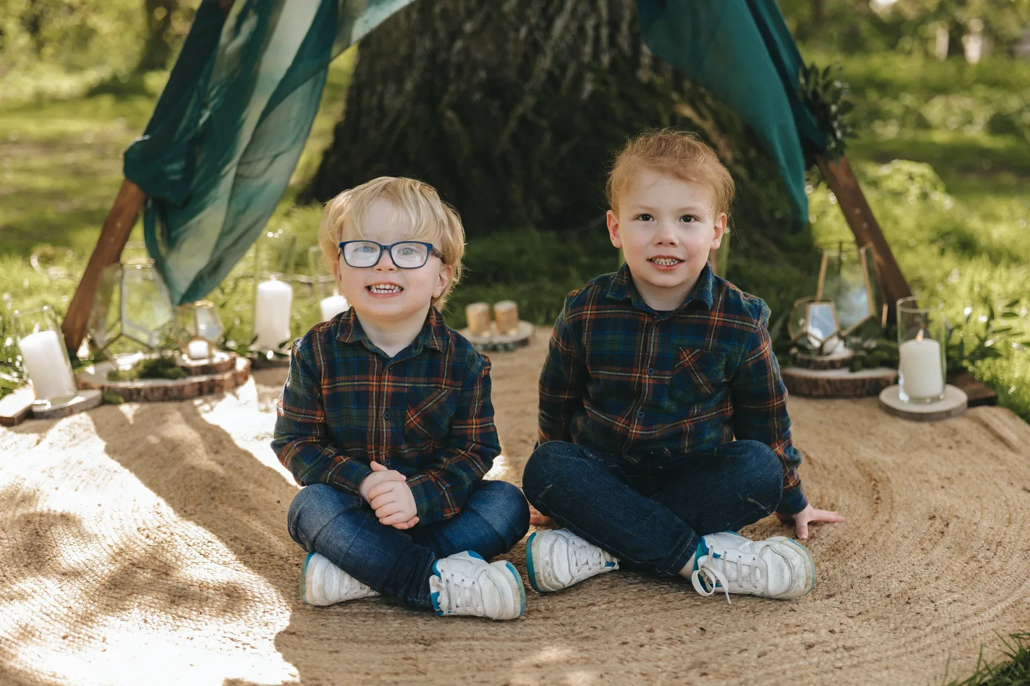 Two young boys wearing plaid shirts and jeans sit on a blanket in a park under a tent made of green fabric draped over a tree. the setting includes grass with scattered candles around them, conveying a cozy outdoor atmosphere.