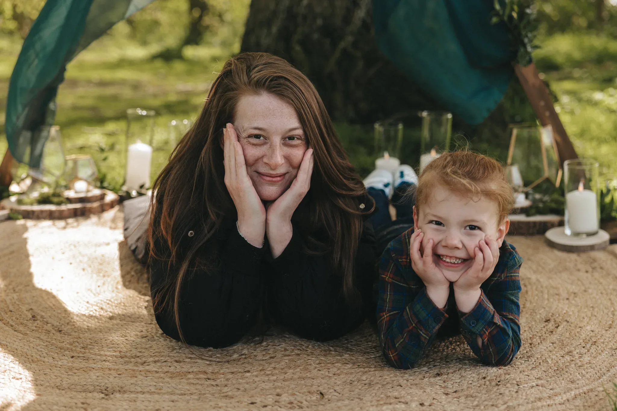 A woman and a young boy lie on their stomachs on a blanket, resting their cheeks on their hands and smiling at the camera, with a picnic setup and a tree draped with teal fabrics in the background.