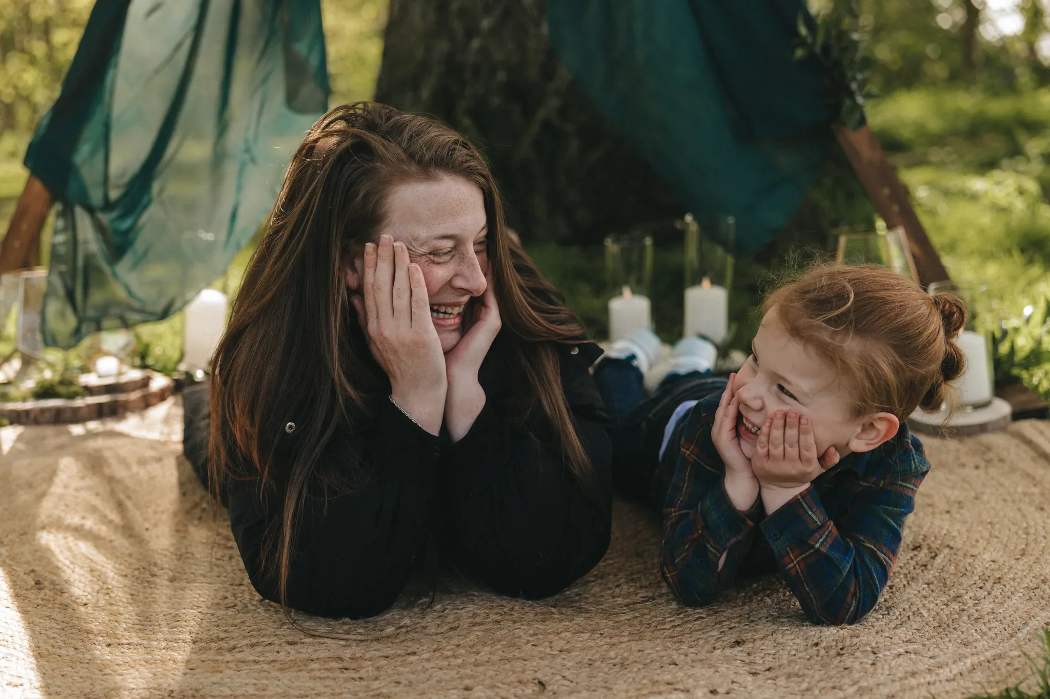 A woman and a young child, both with joyful expressions, lying on a blanket outdoors. the child, wearing a plaid shirt, touches their face mirroring the woman’s pose. greenery and candles in the background create a cozy atmosphere.