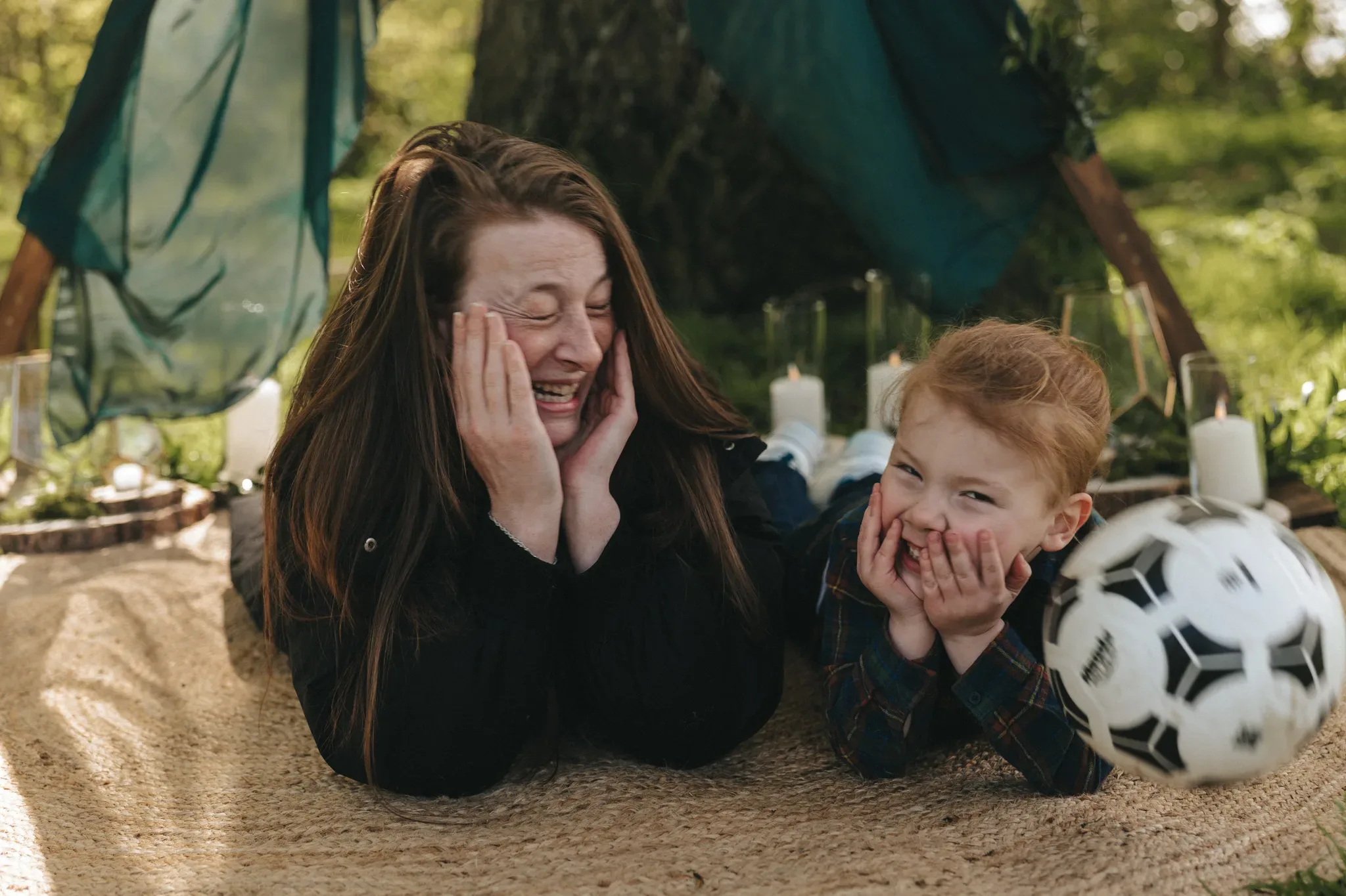 A woman and a young boy laugh joyously while lying on a blanket outdoors, with a soccer ball nearby. both are holding their faces in delight, surrounded by a green, sunlit park setting with soft focus in the background.