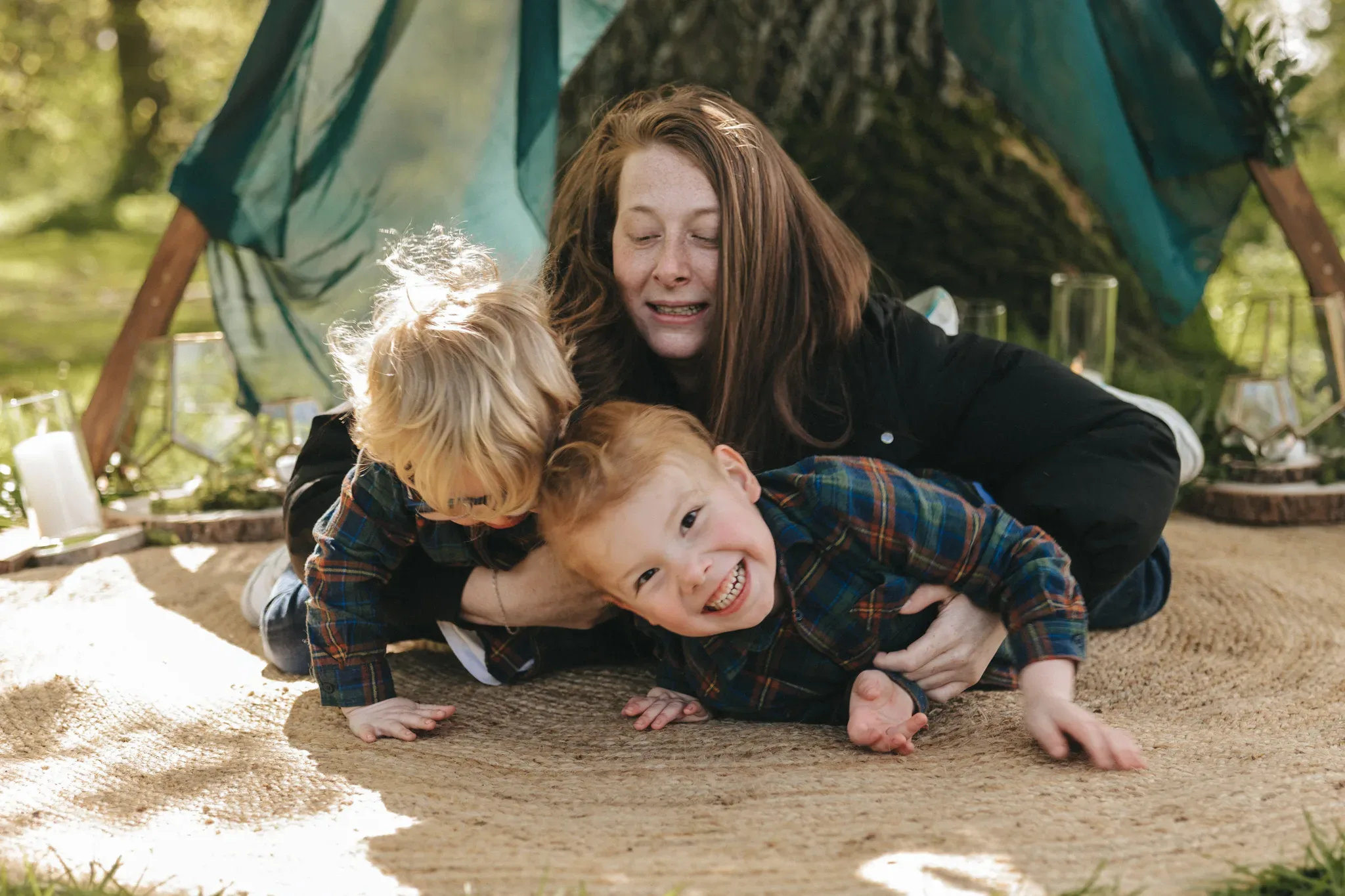 A joyful woman playing with two young boys in a plaid shirt under a makeshift tent outdoors. they're smiling, with sunlight filtering through trees in the background.