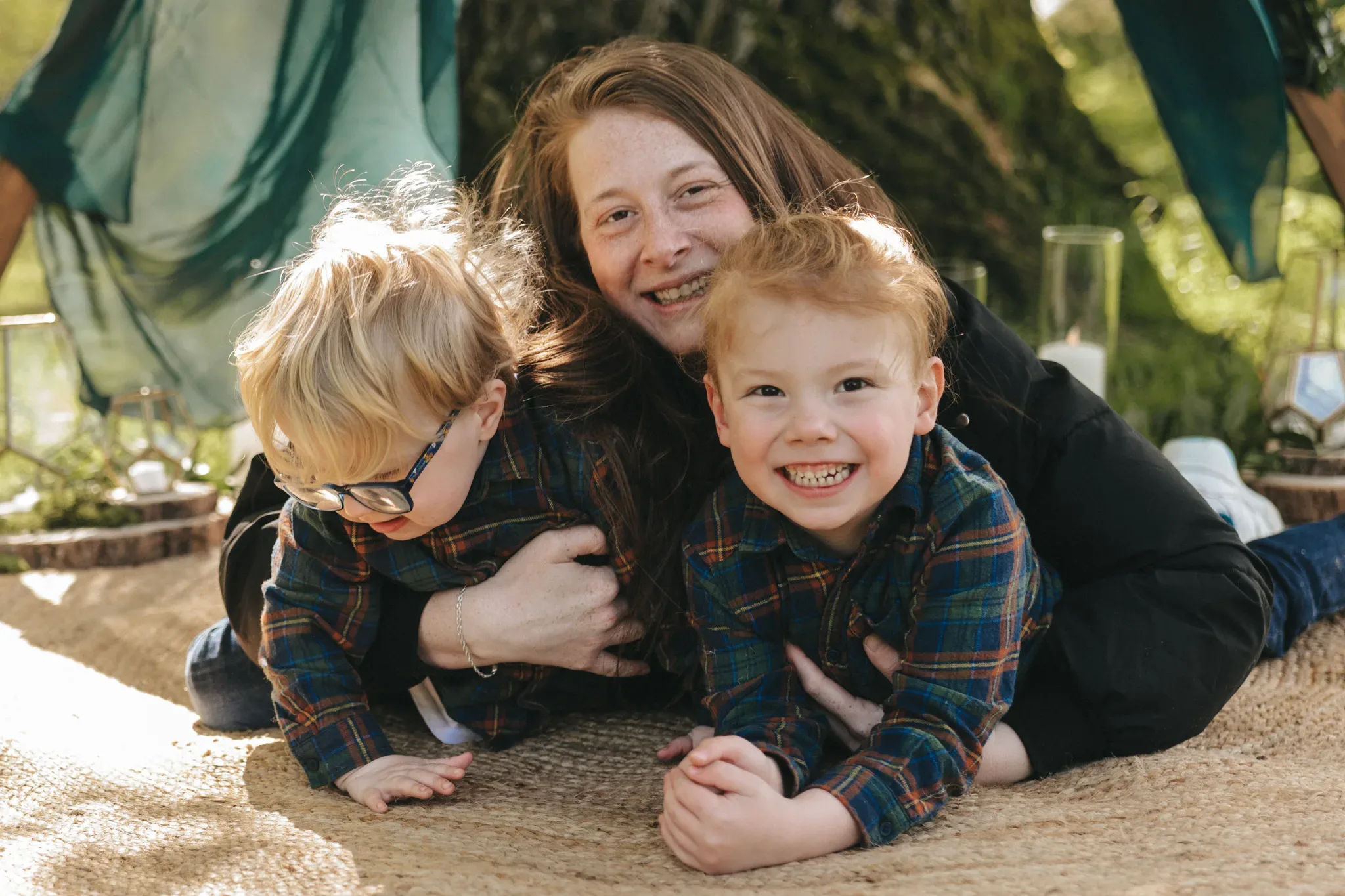 A joyful woman lies on a blanket with her two young sons, all wearing plaid shirts and smiling warmly in a sunlit garden setting with greenery in the background.