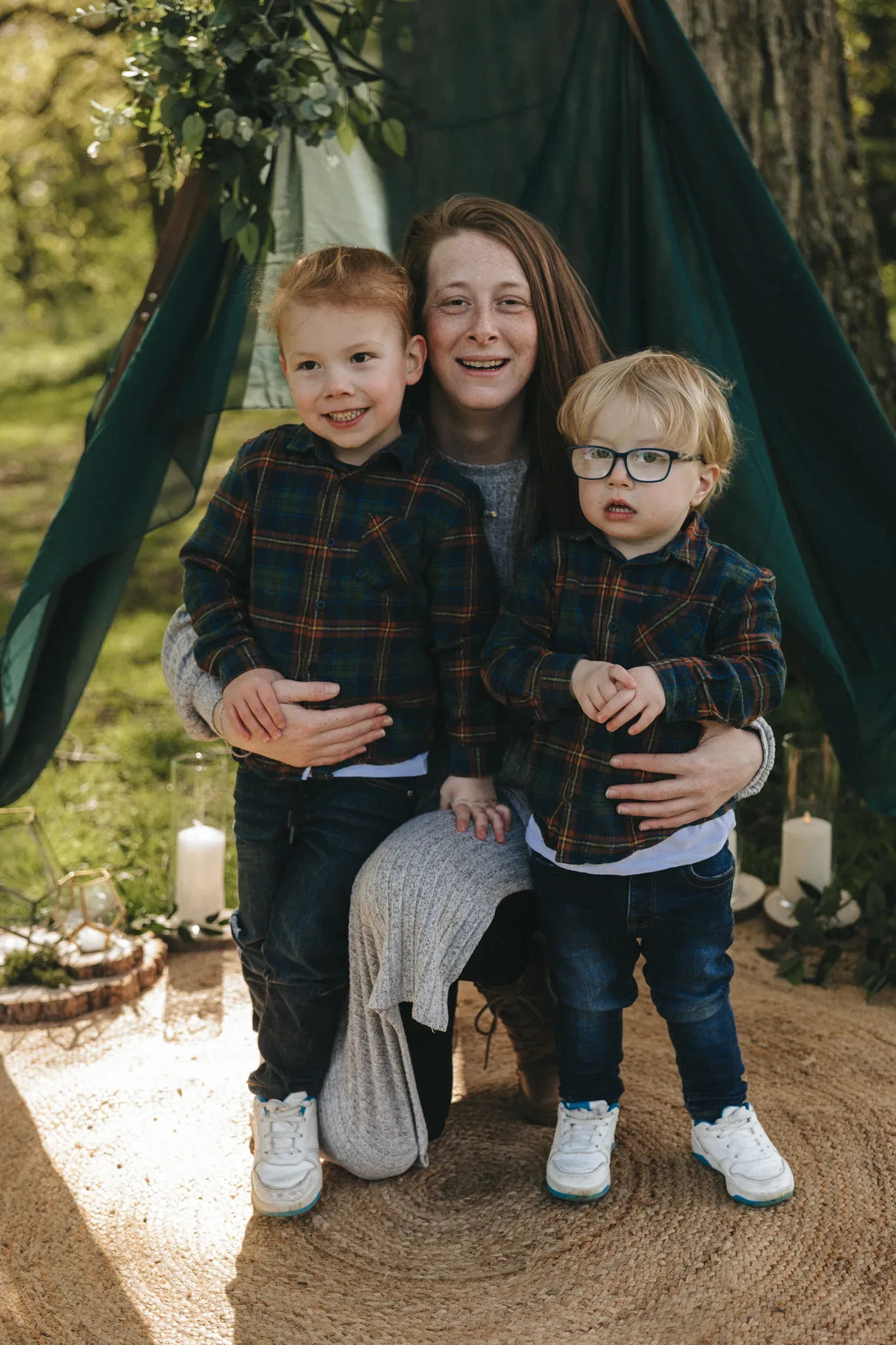A mother sits with her two young sons under a green tent outdoors, smiling. they all wear matching plaid shirts and jeans. the setting includes a circular rug and decorative candles, creating a cozy, woodland ambiance.