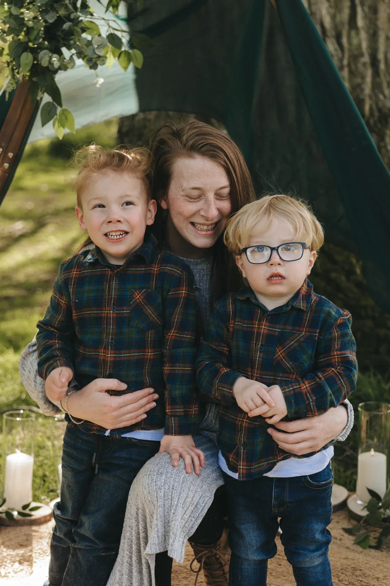 A joyful woman hugged by two young boys in plaid shirts, outdoors under a canopy, smiling brightly with candles and natural light surrounding them.
