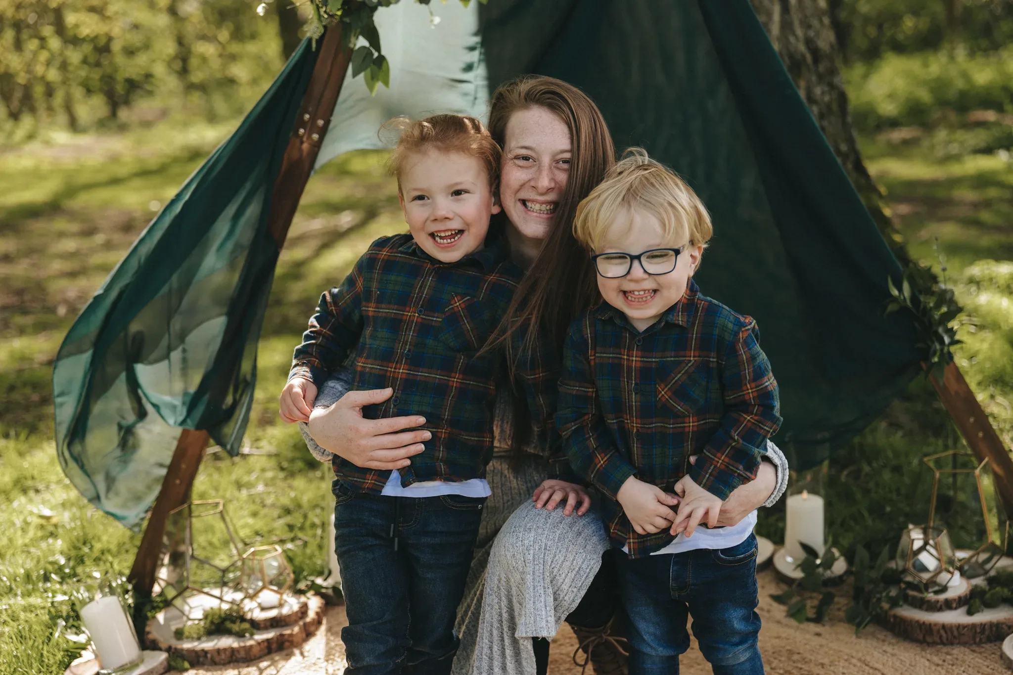 A joyful woman sits outdoors with two young children, all wearing matching plaid shirts, in front of a teepee-style tent adorned with greenery in a sunny, grassy area. the children, one with glasses, smile brightly, holding paper crafts.