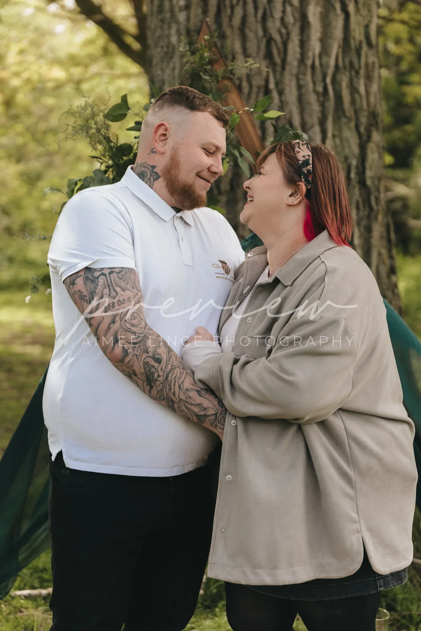 A happy family, a man and a woman named Gabrielle, share a joyful moment outdoors. The man, with tattooed arms, smiles at Gabrielle who has red hair and glasses; they stand