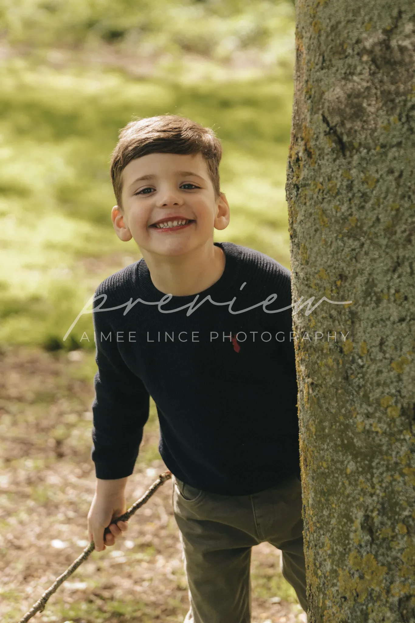 A young boy with short brown hair and a playful smile peeks out from behind a tree. he holds a stick and wears a dark sweater and olive pants. the background is a softly blurred green landscape. the image has a watermark "premien aimee lance photography.
