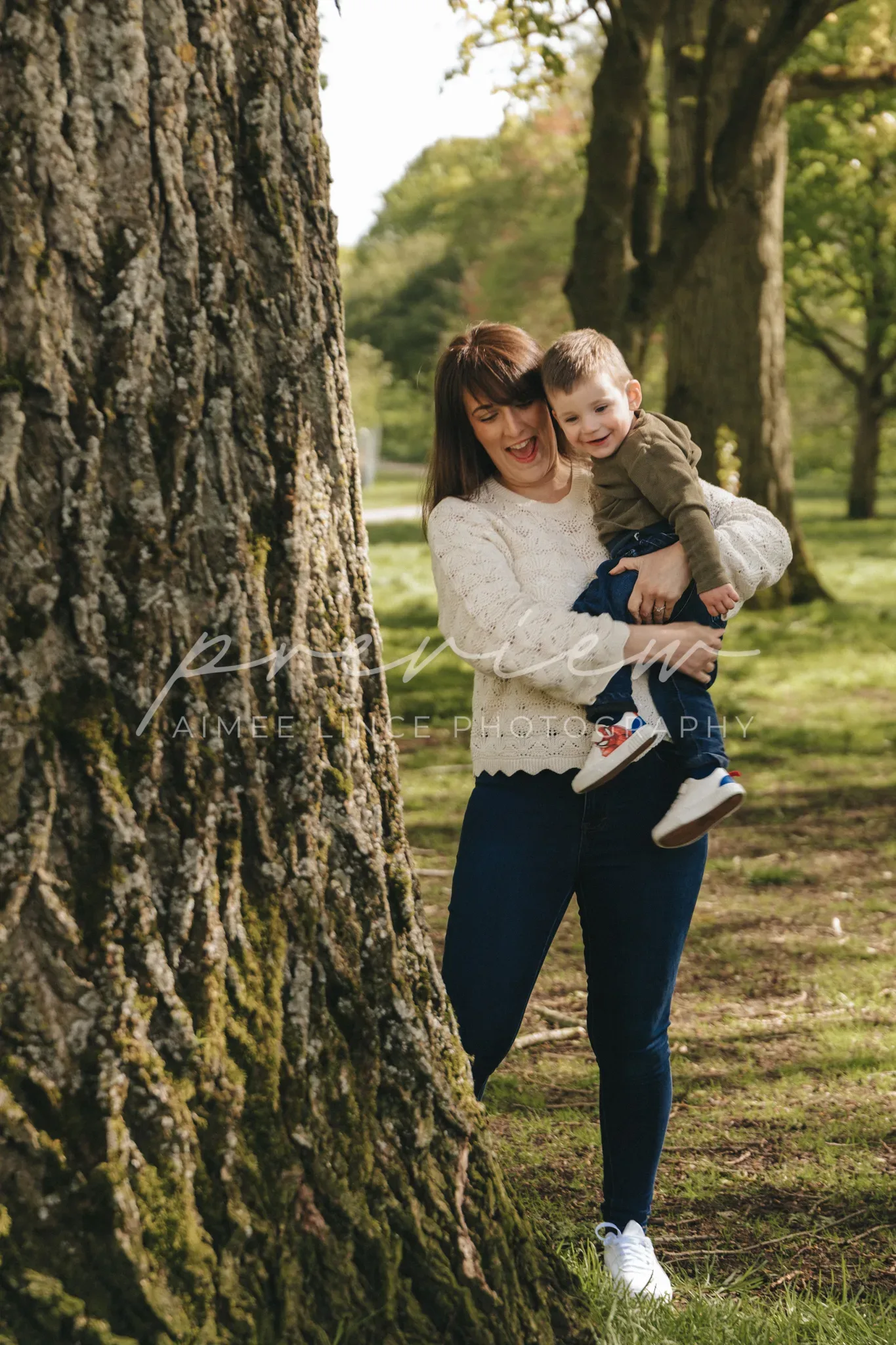A woman in a black top and blue jeans holds a young boy in a sweater and jeans next to a large tree in a sunlit park, both smiling joyfully. the watermark "aimee l. photography" appears on the image.