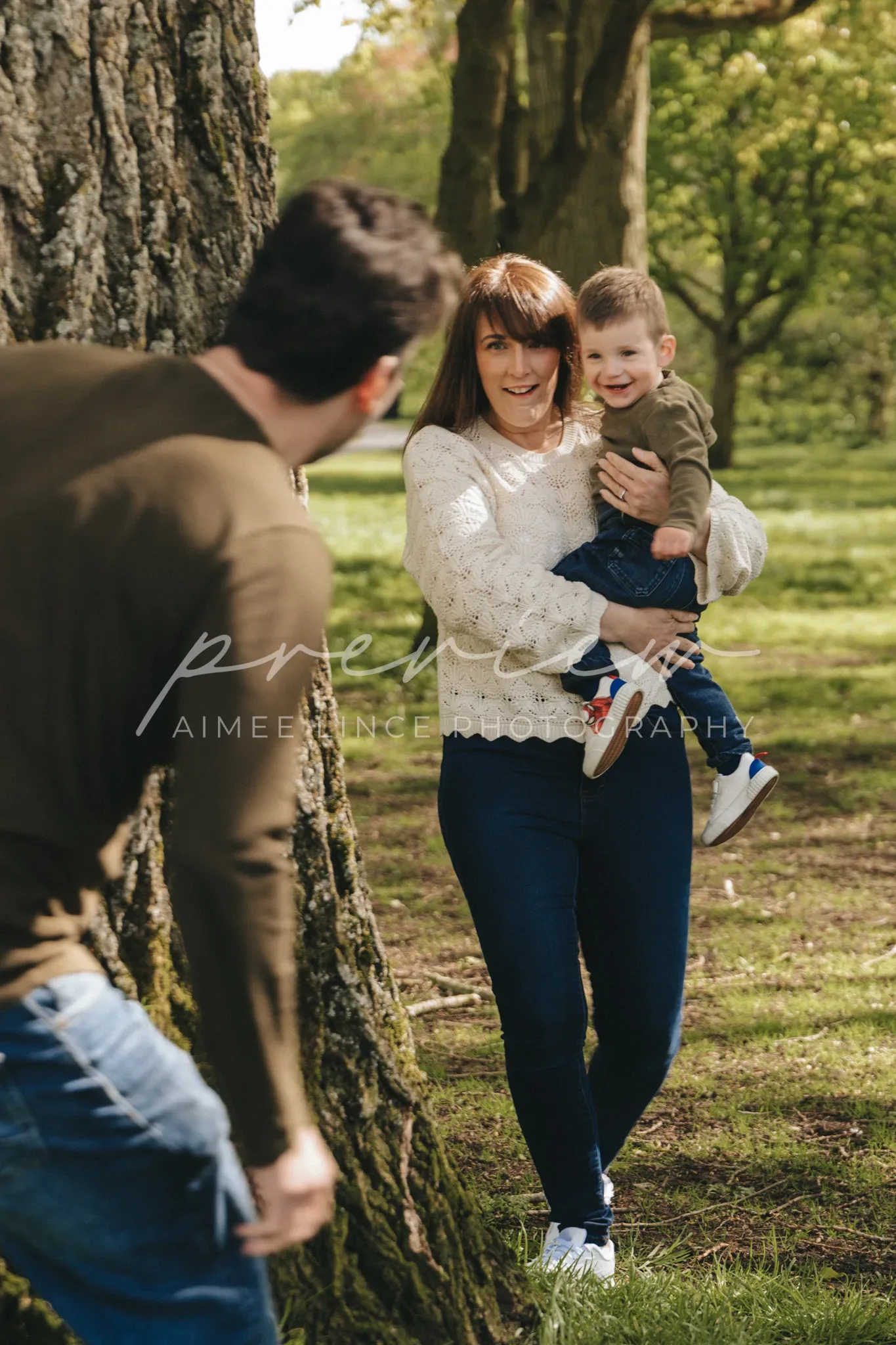 A woman holding a toddler stands smiling near a tree in a park, looking at a man partially visible on the left. the setting is sunny and verdant. both are dressed in casual attire. the mother and child are joyous and engaging with the man.