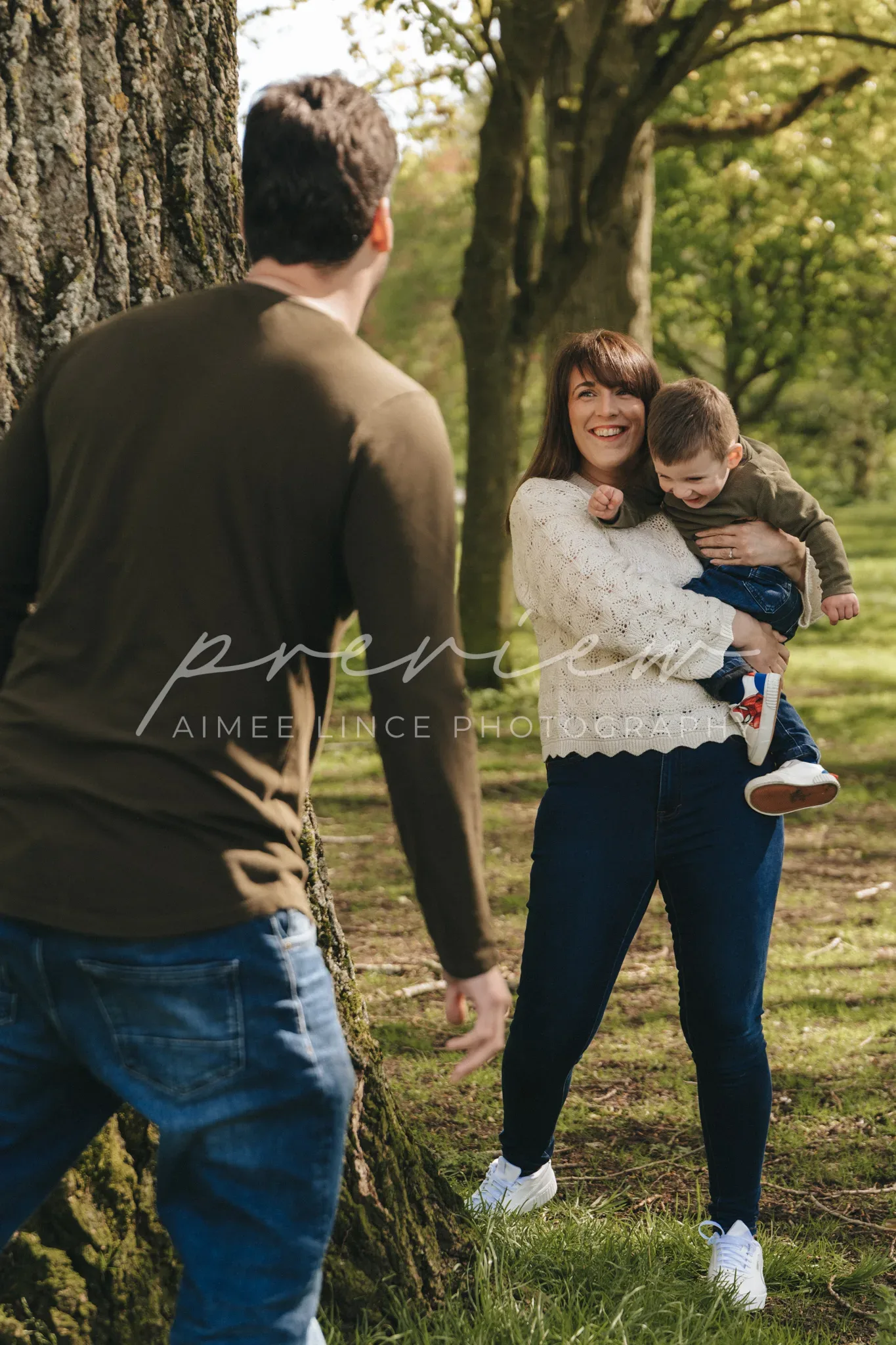 A woman holding a toddler smiles joyously at a man standing foreground in a lush park. the child playfully reaches out toward the man. the scene is bright with sunlight filtering through trees.