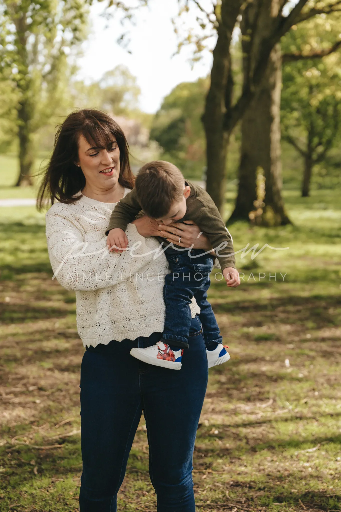 A joyful young woman holds a toddler in a park. the child, wearing a green jacket and jeans, playfully leans forward as the woman, dressed in a white sweater and blue jeans, supports him, both smiling amid sunlit trees.
