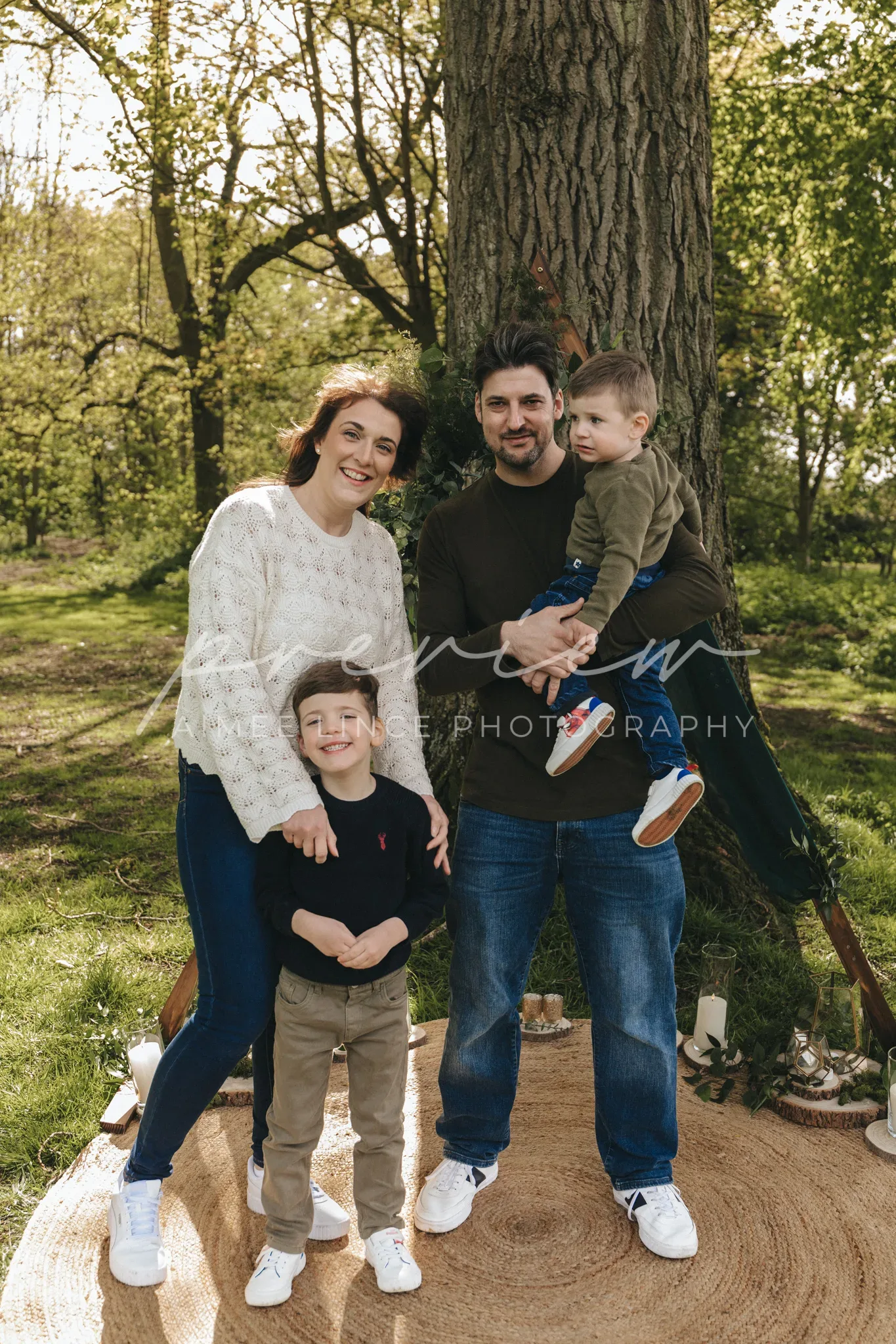 A happy family of four posing outdoors in a park by a large tree. the mother, father, and two young sons all smile at the camera, with one son held in the father's arms and the other standing in front.