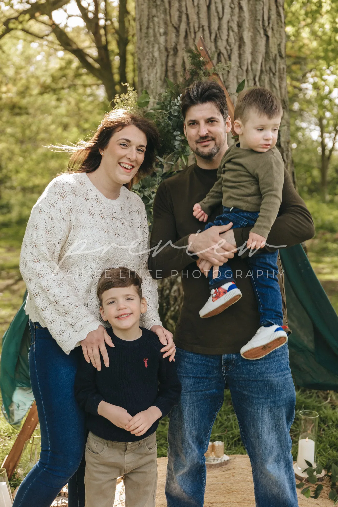 A happy family of four posing outdoors in a park. the mother and father are standing with their young son between them; the father holds a toddler on his hip. all are smiling, surrounded by lush greenery.