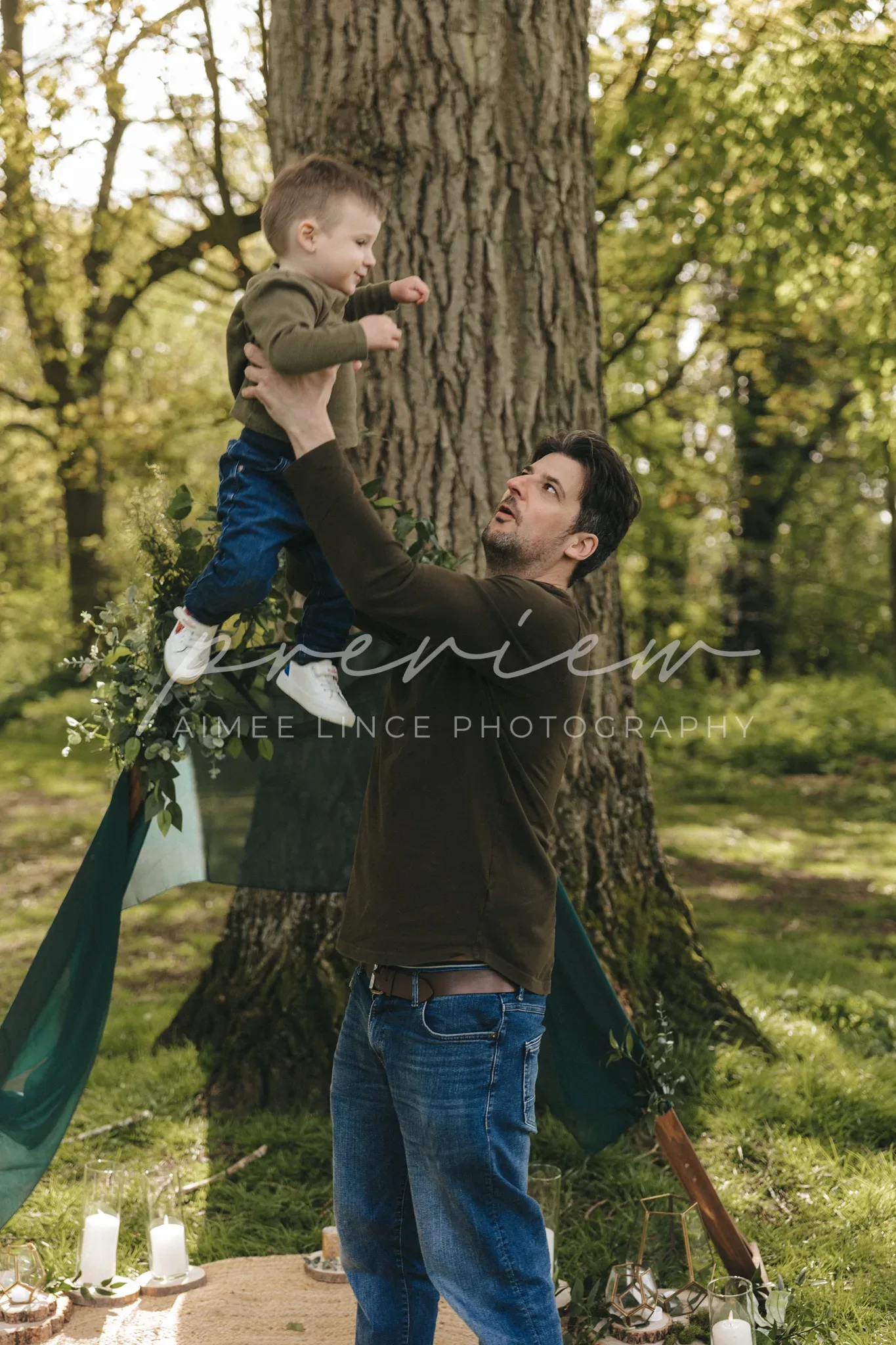 A man lifts a toddler high in his arms outdoors, both smiling, amidst large trees and soft sunlight. the setting includes some candles at their feet, adding a warm ambiance to the scene.