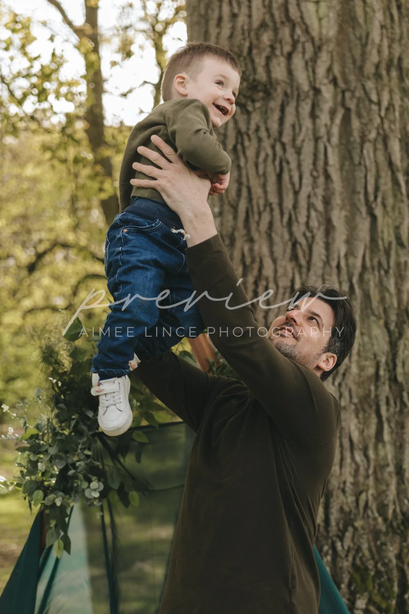 A joyful man outdoors lifts a laughing toddler high in the air against the backdrop of a large tree. both are dressed in casual long-sleeved tops, sharing a moment of happiness in a sunlit park.