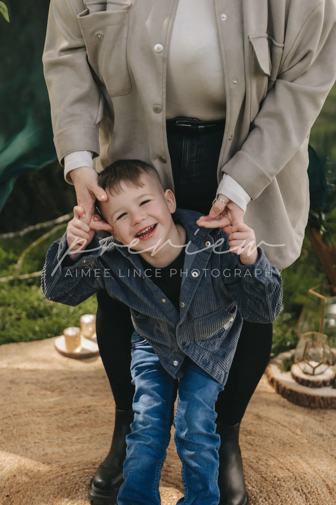 A joyful toddler boy, Gabrielle, smiles brightly, held playfully by his mother's hands around his ears. He wears a denim jacket and jeans. The backdrop features lush greenery, soft lighting