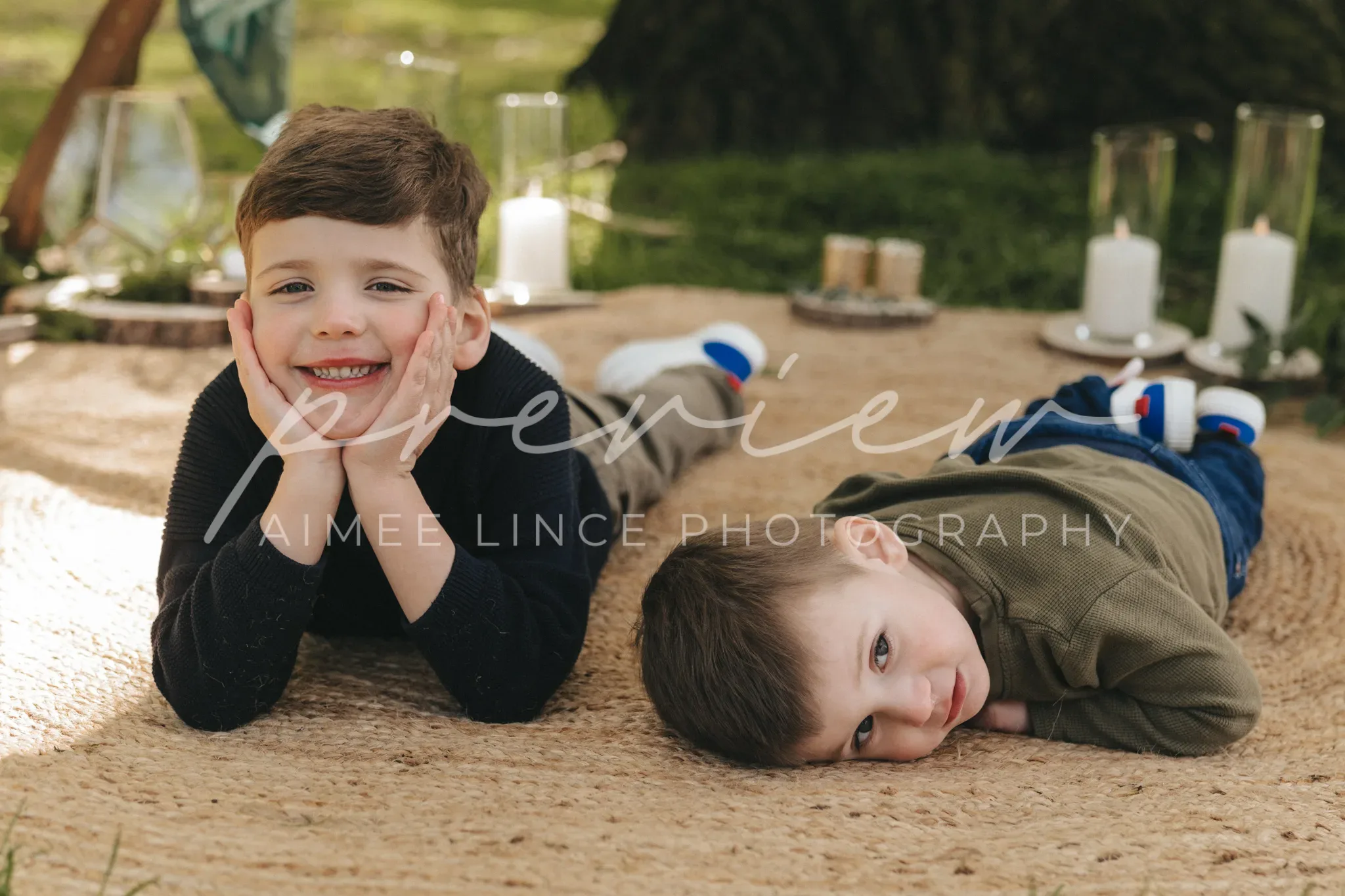 Two young boys lying on a blanket in a park, smiling at the camera. one boy has his chin resting on his hands, and the other boy is laying his head on the blanket, cheek pressed down. candles and lanterns are around them.