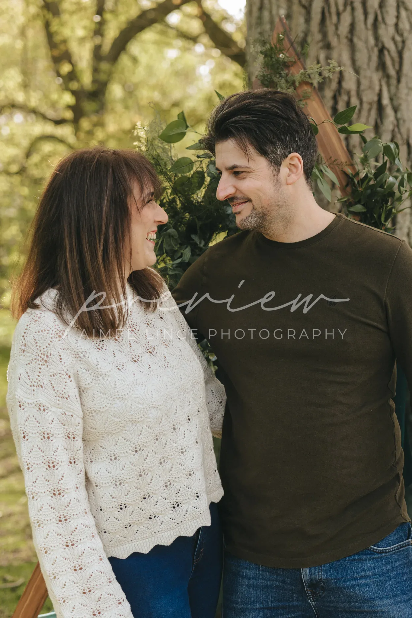 A man and a woman smile at each other, standing close under a tree. the woman wears a white knitted sweater and jeans, and the man wears a green shirt and jeans. the background is a lush park with soft sunlight filtering through the leaves.