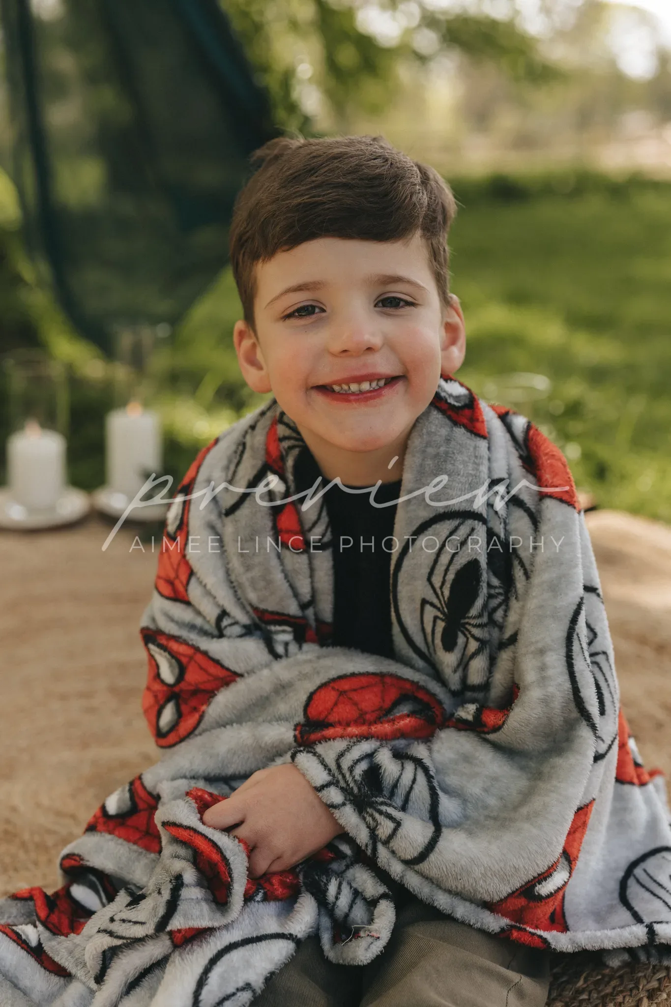 A young boy with short dark hair, smiling, wrapped in a mickey mouse blanket, sitting outdoors. the background features soft-focused trees and subtle sunlight, enhancing the tranquil setting.