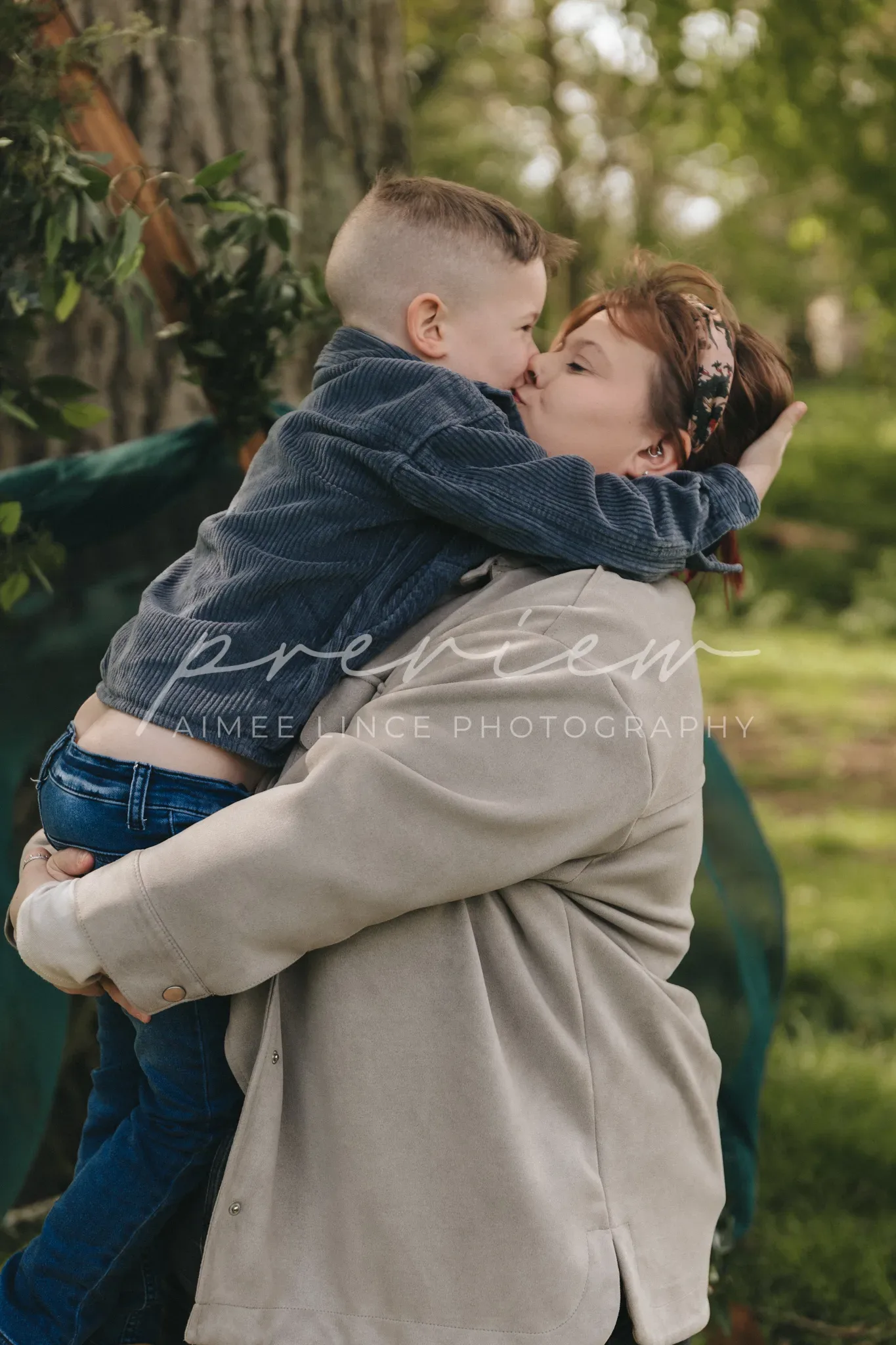 A young woman named Gabrielle, carrying a small boy in her arms, sharing a kiss on the lips in a lush park. Gabrielle's right arm encircles the boy's back, both showing