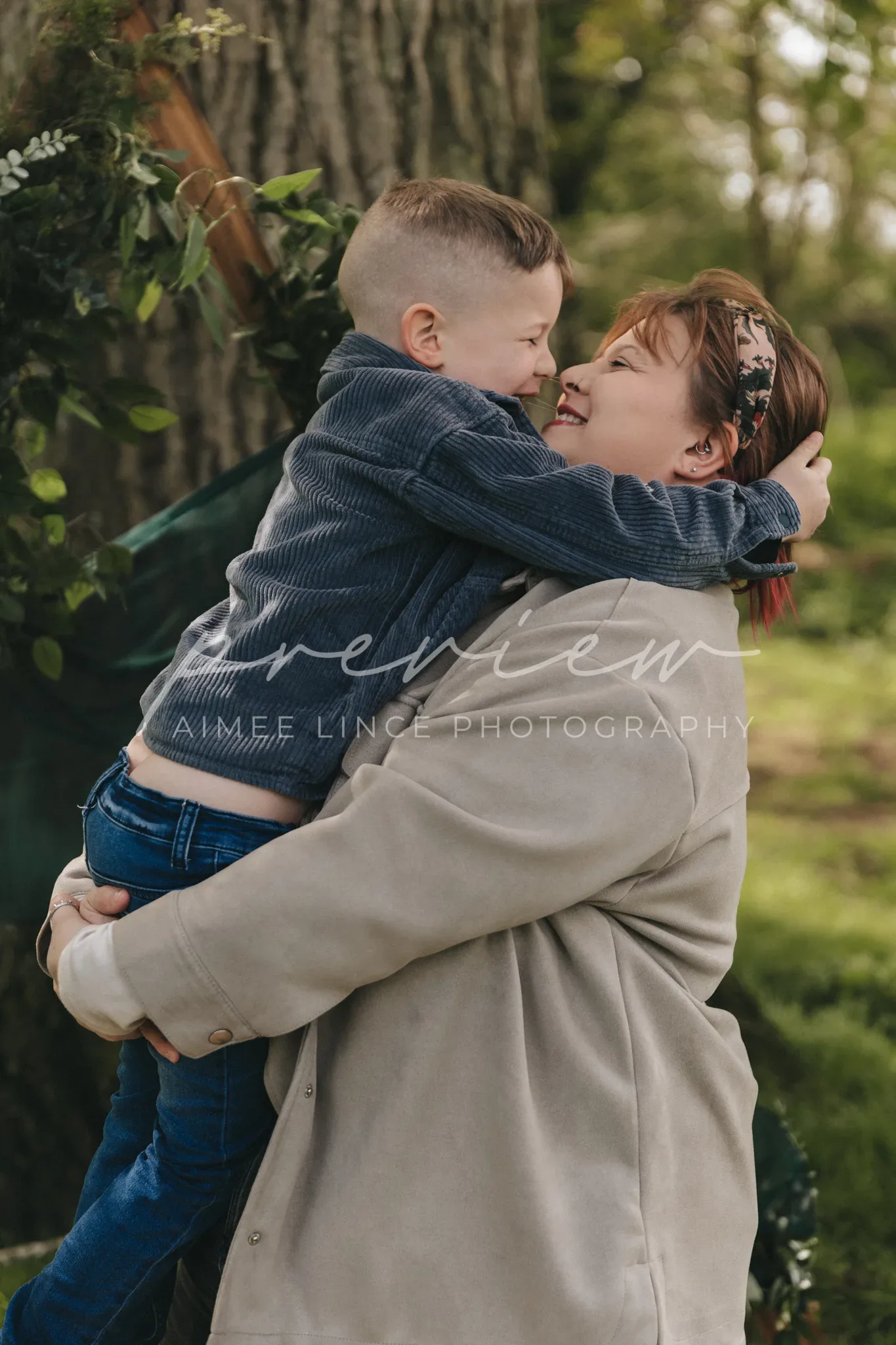 A woman in a beige coat hugs a young boy wearing denim. They smile lovingly at each other, nose-to-nose, in a lush park setting. The watermark "Gabrielle Family Photography