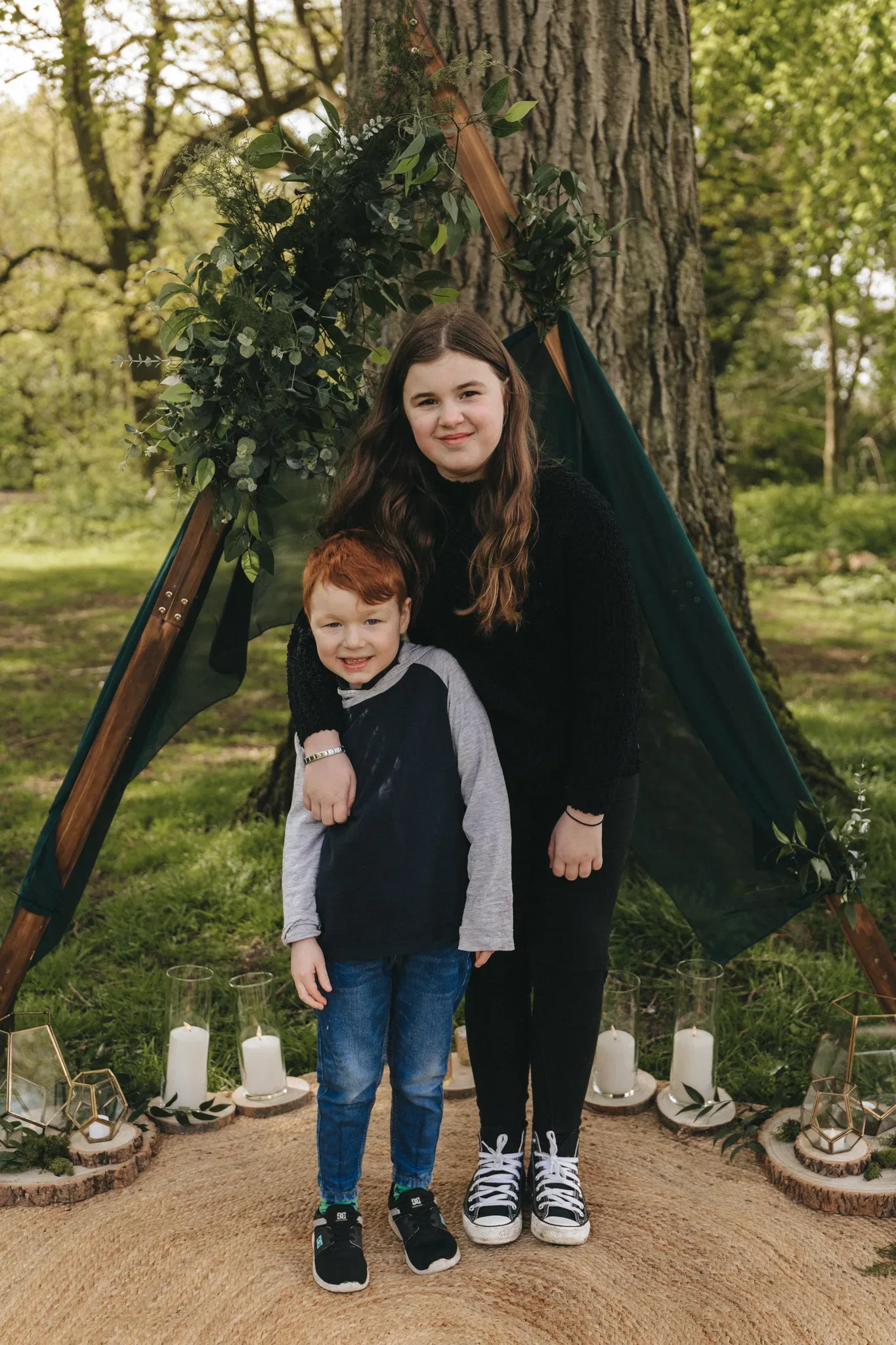 A young girl with long brown hair and a black sweater stands beside a younger boy with red hair and a blue vest, both smiling in front of a wooden teepee adorned with greenery, set in a lush park.