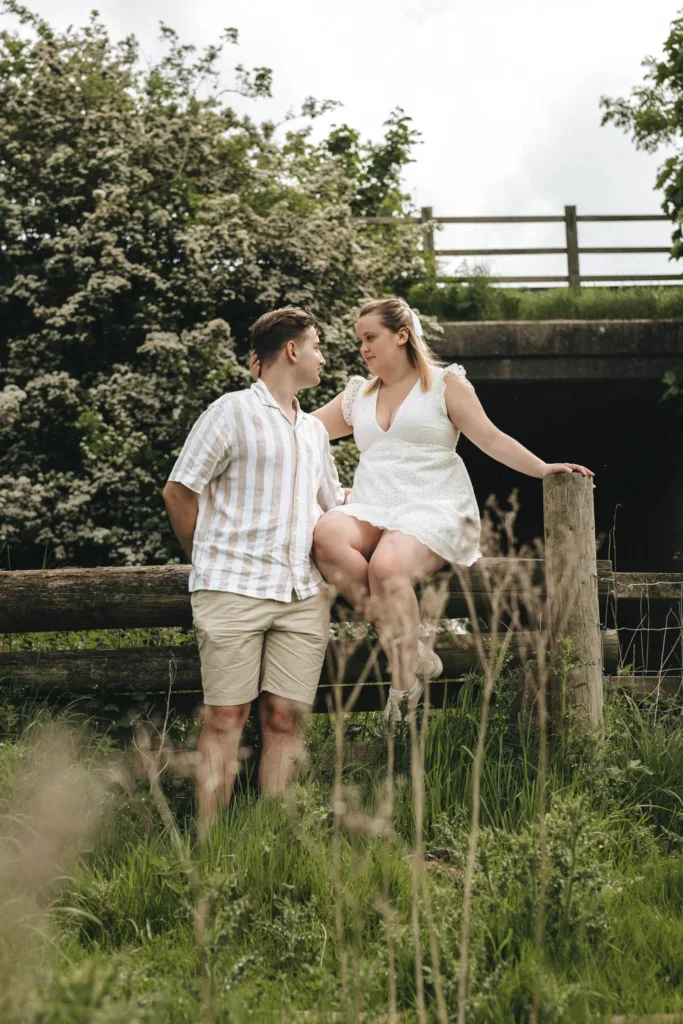 A couple outdoors, set against the serene Irby Dales Wood backdrop. The woman, in a white dress, sits playfully on a wooden fence post. The man stands beside her in a striped shirt and shorts. Both appear engaged in conversation, surrounded by lush greenery typical of Lincolnshire near Grimsby. © Aimee Lince Photography - Wedding photographer in Lincolnshire, Yorkshire & Nottinghamshire