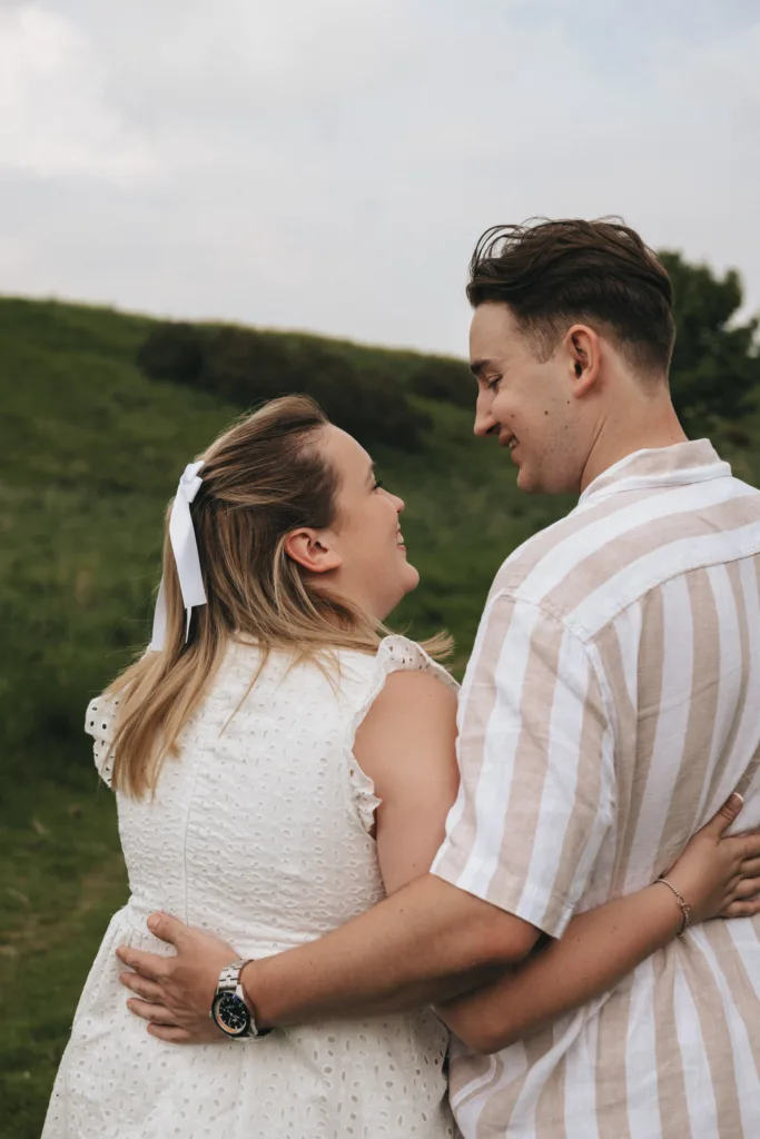 On a grassy hill in Irby Dales Wood, a couple stands closely, embracing each other. The woman, in a white dress with a ribbon in her hair, smiles up at the man wearing a short-sleeve striped shirt. Under the cloudy sky, their affection and sense of togetherness hint at an engagement to cherish. © Aimee Lince Photography - Wedding photographer in Lincolnshire, Yorkshire & Nottinghamshire