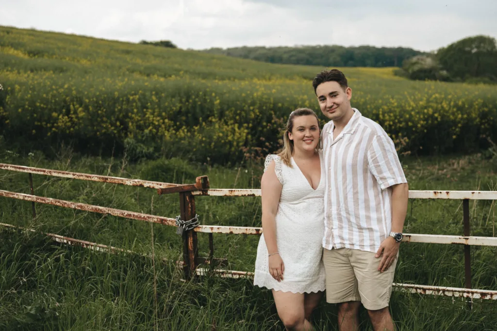 A couple celebrates their engagement, smiling in front of a rustic metal gate in the lush Irby Dales Wood. The woman wears a white dress, and the man sports a striped shirt and shorts. Yellow flowers bloom across the expansive landscape, with a line of trees under a cloudy sky. © Aimee Lince Photography - Wedding photographer in Lincolnshire, Yorkshire & Nottinghamshire