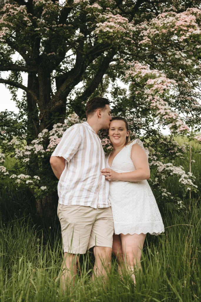 In the serene Irby Dales Wood, a couple stands in tall grass under a tree with pink blossoms. The person on the left, in a striped shirt and khaki shorts, leans in to whisper sweet words of engagement to their smiling partner in a white dress. The lush greenery provides an enchanting backdrop. © Aimee Lince Photography - Wedding photographer in Lincolnshire, Yorkshire & Nottinghamshire