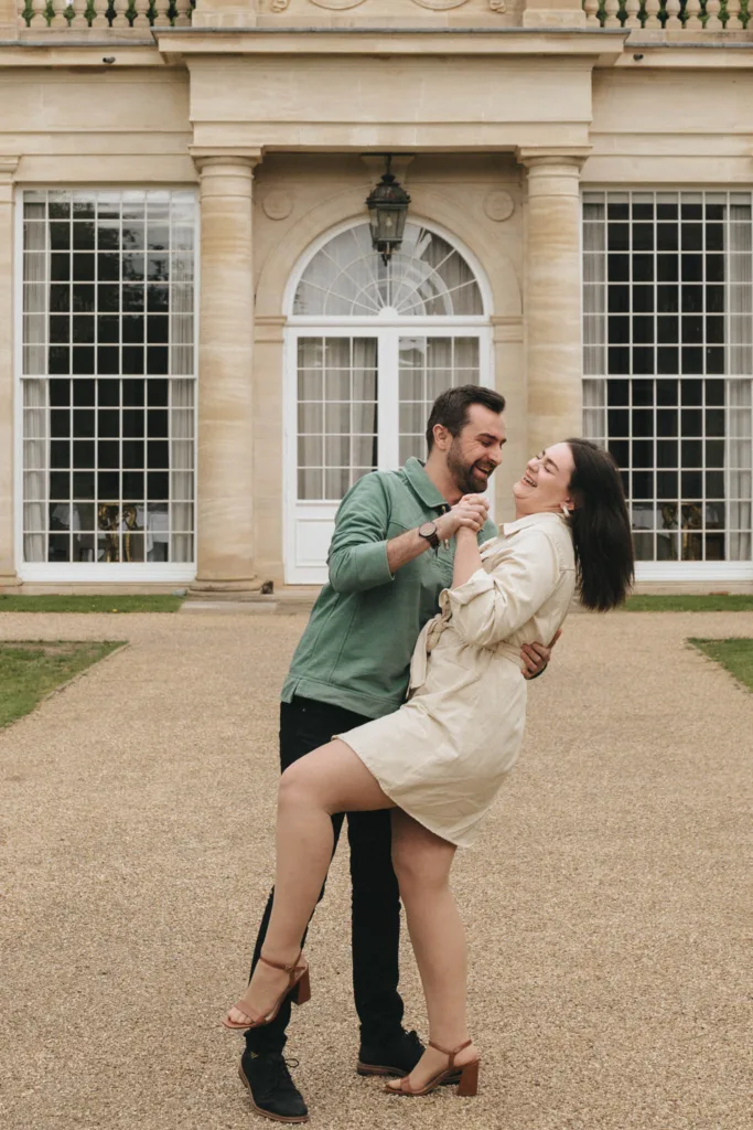 A couple is playfully dancing on a gravel path in front of Rushton Hall's grand facade with tall windows. The man, in a green shirt and black pants, holds the woman's hand. She wears a beige dress, smiling as he dips her gracefully during their elegant outdoor photoshoot near Kettering. © Aimee Lince Photography - Wedding photographer in Lincolnshire, Yorkshire & Nottinghamshire