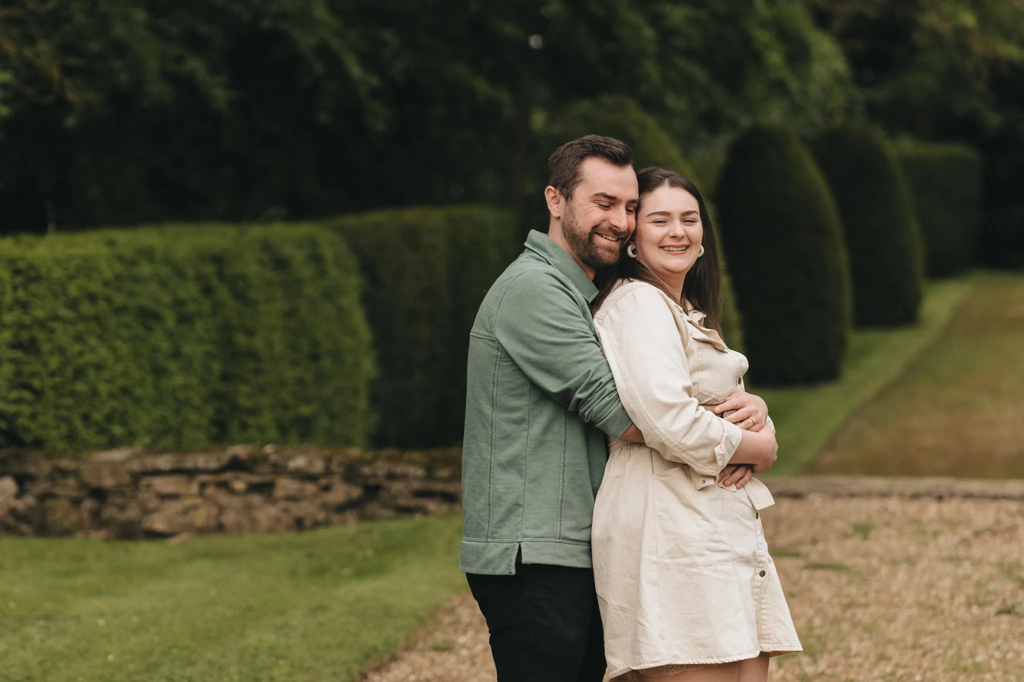 A couple stands embracing in a lush green park, perfect for a romantic photoshoot. The man, wearing a green jacket at Rushton Hall near Kettering, smiles as he hugs the woman from behind. She, in a light-colored dress, smiles with closed eyes against the serene backdrop of hedges and grass pathways. © Aimee Lince Photography - Wedding photographer in Lincolnshire, Yorkshire & Nottinghamshire