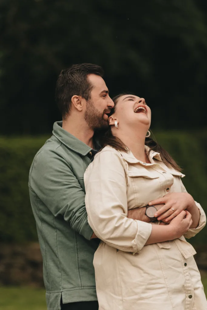A couple embraces joyfully outdoors in Kettering. The man in a green jacket hugs the woman from behind as she laughs heartily, leaning back against him during their engagement photoshoot. They are surrounded by greenery, suggesting a peaceful, natural setting. © Aimee Lince Photography - Wedding photographer in Lincolnshire, Yorkshire & Nottinghamshire