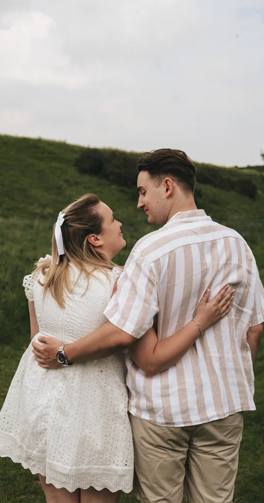 On a grassy hill near Irby Dales Wood, a couple stands embracing, gazing into each other's eyes. The woman in a white dress with a bow in her hair and the man in a striped shirt and khaki pants create an idyllic scene against the overcast sky, capturing the essence of their engagement. © Aimee Lince Photography - Wedding photographer in Lincolnshire, Yorkshire & Nottinghamshire