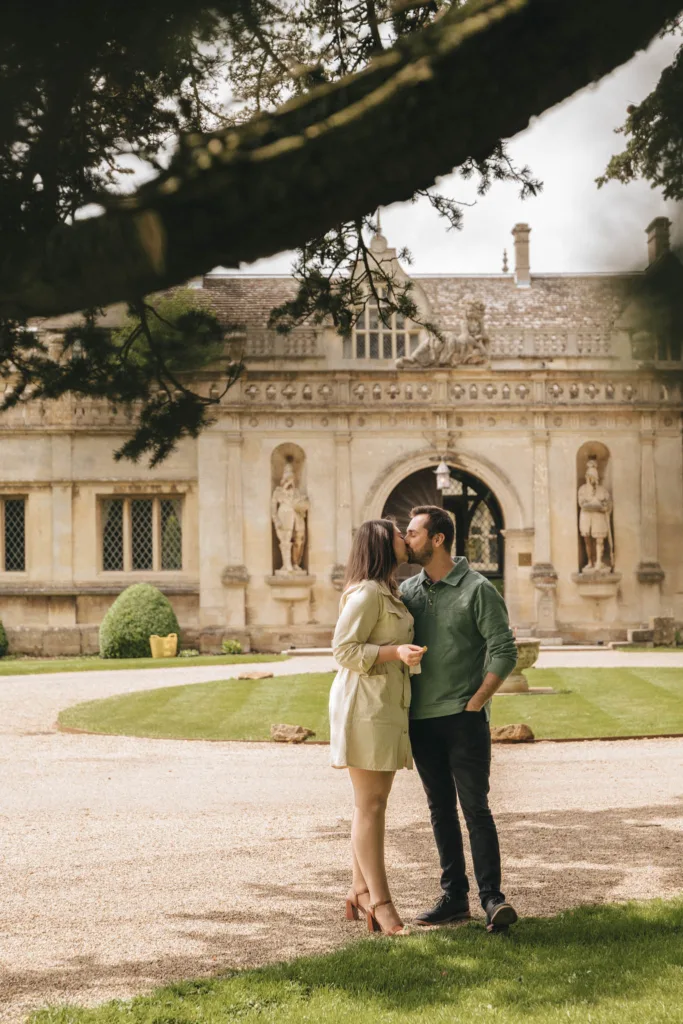 A couple stands on a gravel path in front of an old stone building with statues at Rushton Hall. The woman, in a light dress and heels, leans in towards the man during their engagement photoshoot. A large tree branch arcs above them, and manicured lawns surround the scene. © Aimee Lince Photography - Wedding photographer in Lincolnshire, Yorkshire & Nottinghamshire