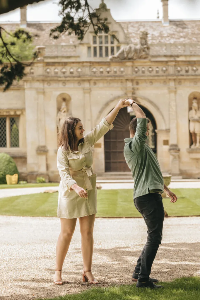 A couple dances joyfully on a gravel path in front of an ornate, historical building during their engagement photoshoot. Stone statues and large wooden doors stand behind them as the woman, in a light-colored dress and heels, twirls with the man in a green shirt. Lush greenery surrounds them. © Aimee Lince Photography - Wedding photographer in Lincolnshire, Yorkshire & Nottinghamshire