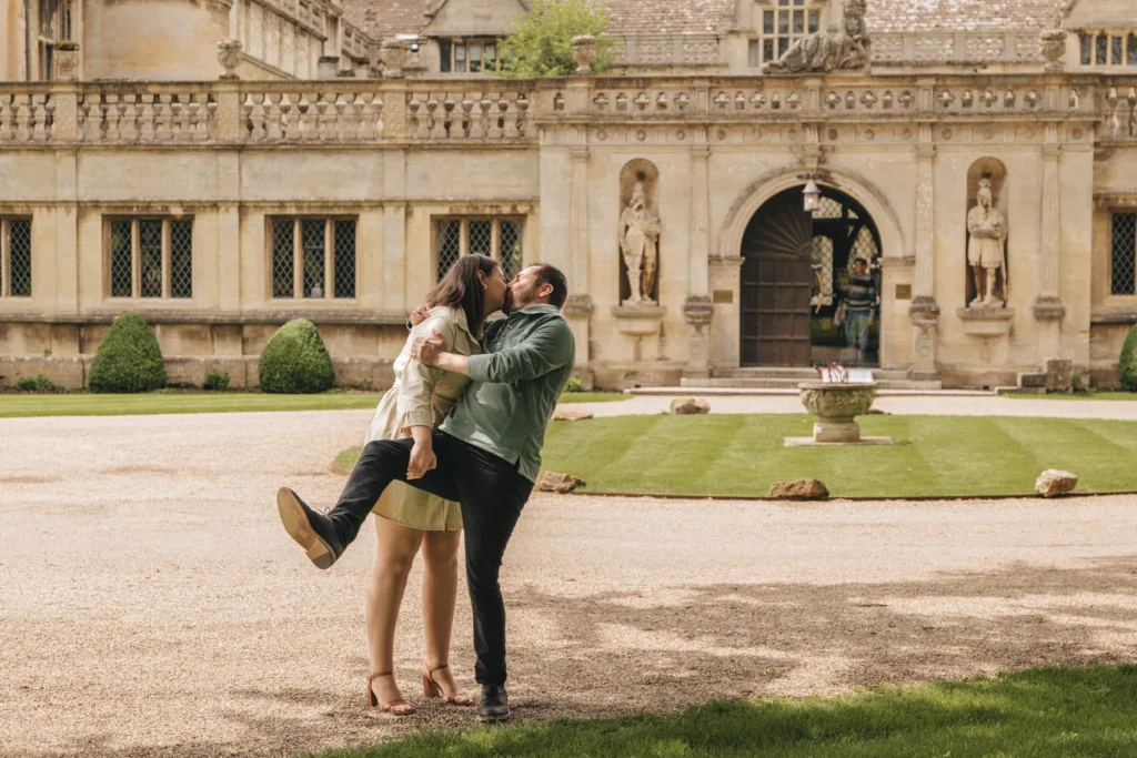 A couple shares a joyful embrace on the grounds of Rushton Hall, an elegant historic building near Kettering, renowned for its ornate architecture and statues. The playful atmosphere is enhanced as the man lifts the woman, with well-maintained lawns and a central stone urn under a partly cloudy sky adding to the garden's charm. © Aimee Lince Photography - Wedding photographer in Lincolnshire, Yorkshire & Nottinghamshire