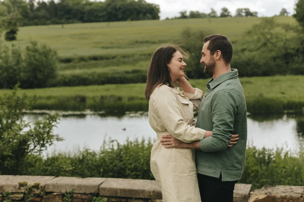 A couple stands by a stone wall at Rushton Hall, embracing and smiling at each other during their engagement photoshoot. The woman wears a beige dress, and the man sports a green jacket. A serene pond, lush green fields, and distant trees under a cloudy sky enhance the tranquil setting. © Aimee Lince Photography - Wedding photographer in Lincolnshire, Yorkshire & Nottinghamshire