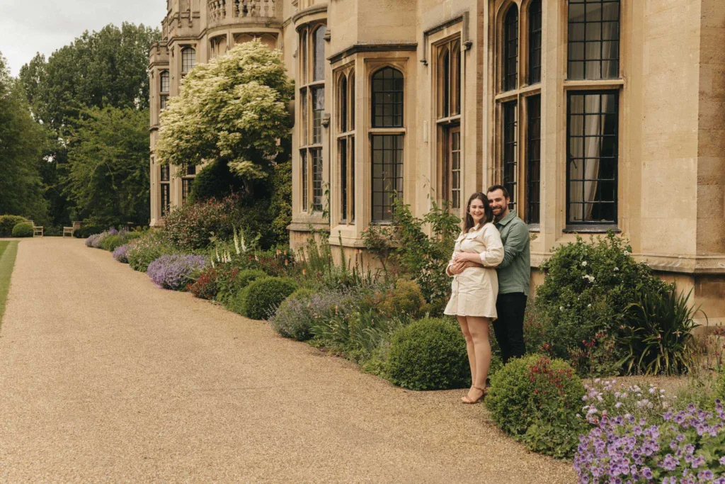A couple stands close on a gravel path beside the historic Rushton Hall with its tall windows. Surrounded by lush greenery and colorful flowers, she wears a light dress, and he sports a green shirt. The setting is peaceful and elegant, perfect for an engagement moment amid manicured gardens near Kettering. © Aimee Lince Photography - Wedding photographer in Lincolnshire, Yorkshire & Nottinghamshire