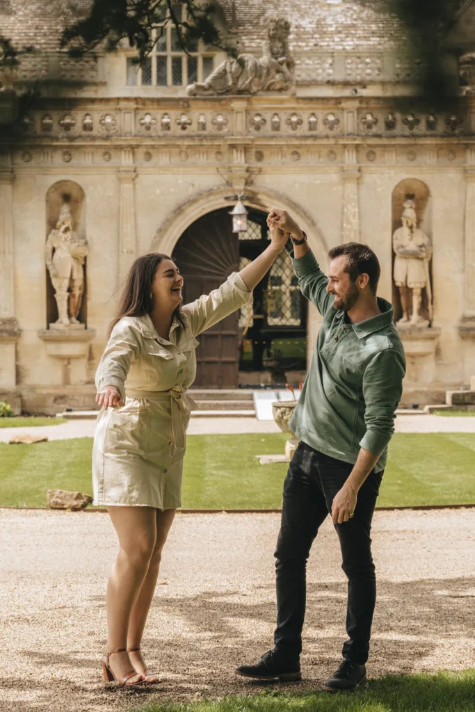 A couple dances joyfully outside the historic Rushton Hall, with statues and an ornate doorway. The woman, in a light dress, spins with one arm raised by the man in a green sweater and jeans. They are surrounded by manicured grass and gravel, capturing their joyful moment under the sun—perfect for a photoshoot. © Aimee Lince Photography - Wedding photographer in Lincolnshire, Yorkshire & Nottinghamshire