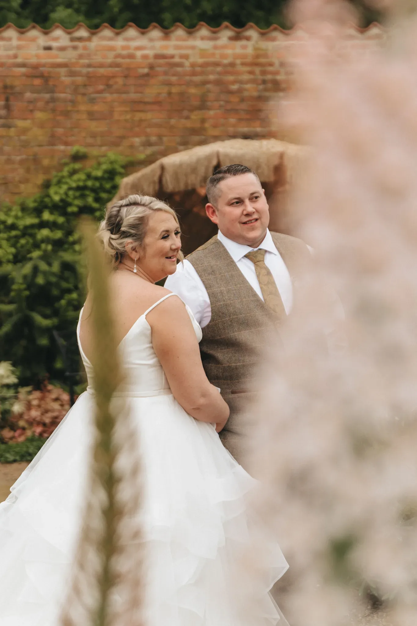 Natalie & Will stand together outdoors, facing slightly to the right. Natalie, in a white off-shoulder wedding dress, looks joyful beside Will, who is dressed in a brown vest and beige tie. Blurred foliage in the foreground and a brick wall in the background frame this heartfelt moment.