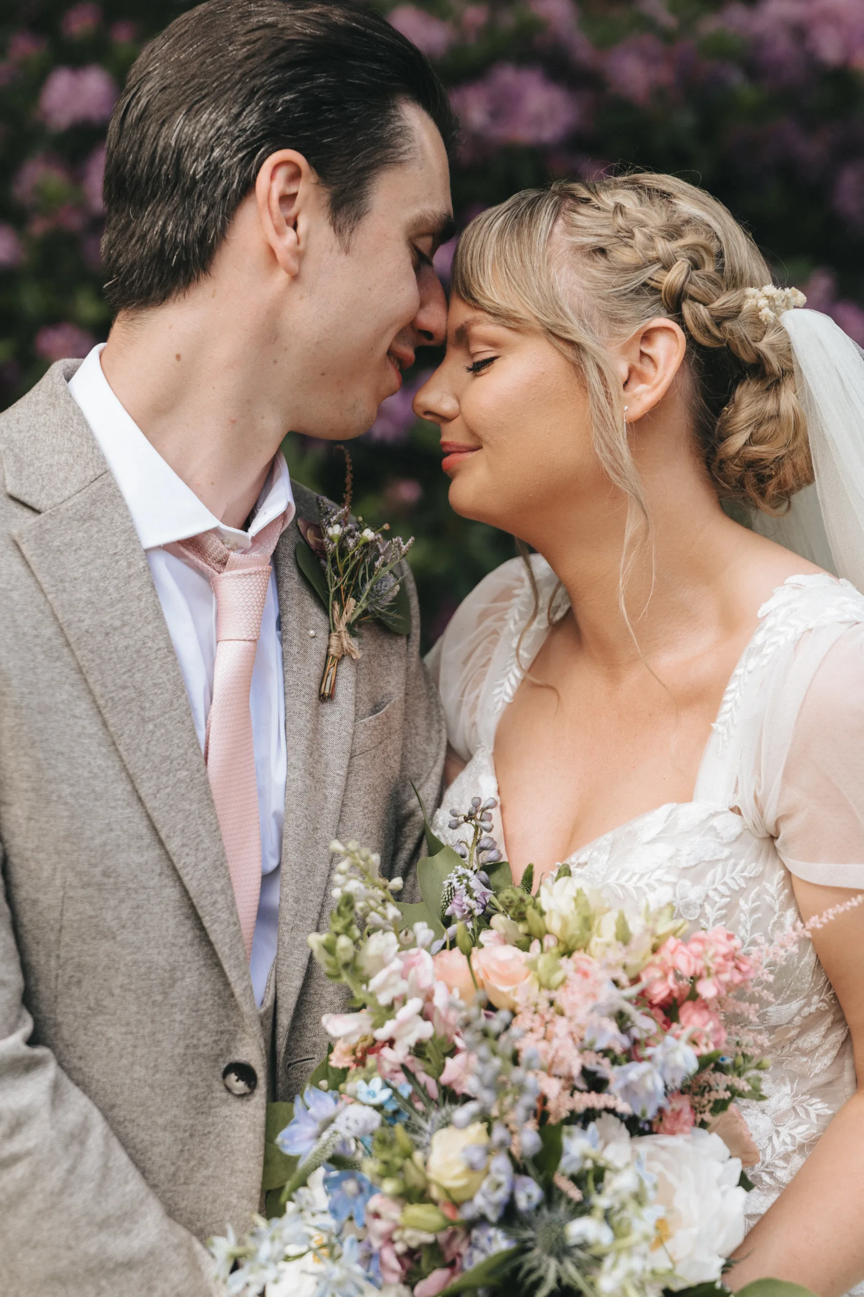 A bride and groom stand closely, touching noses while smiling gently, in front of a backdrop of purple flowers. The groom is dressed in a light gray suit and a pink tie, with a small boutonniere pinned to his lapel. The bride is in a white dress with lace details, holding a vibrant flower bouquet. © Aimee Lince Photography