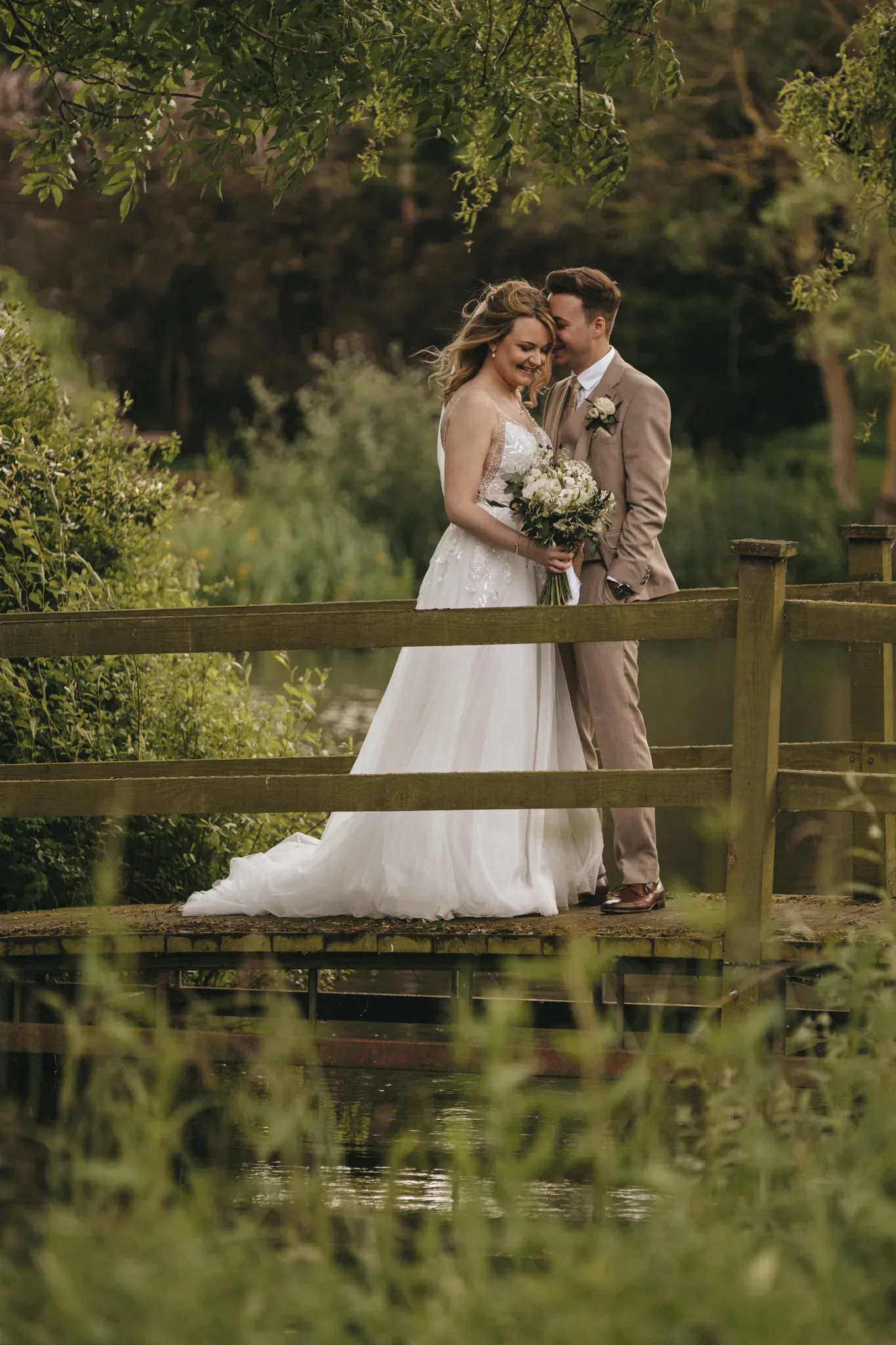 A bride in a white gown and a groom in a beige suit stand on a wooden bridge surrounded by lush greenery. The bride holds a bouquet and smiles, while the groom gently embraces her from behind. The background features trees and a blurred natural setting, creating a serene, romantic atmosphere.