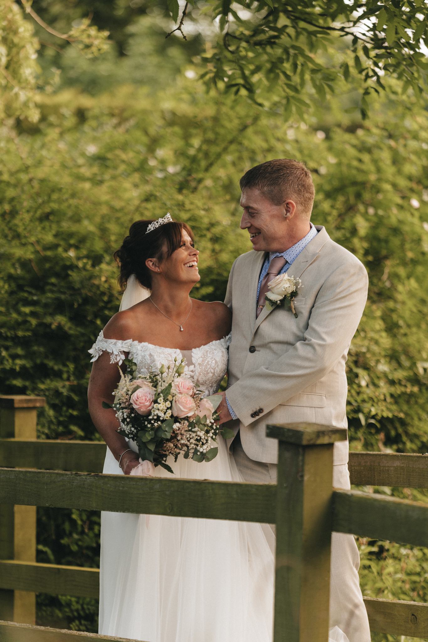 A smiling bride and groom stand close together outdoors at Hall Farm Hotel in Lincolnshire, framed by greenery. The bride wears a white off-the-shoulder gown, a veil, and tiara, holding a bouquet of roses. The groom is in a light-colored suit with a pink tie and boutonniere. They share a joyful moment by the wooden fence. © Aimee Lince Photography